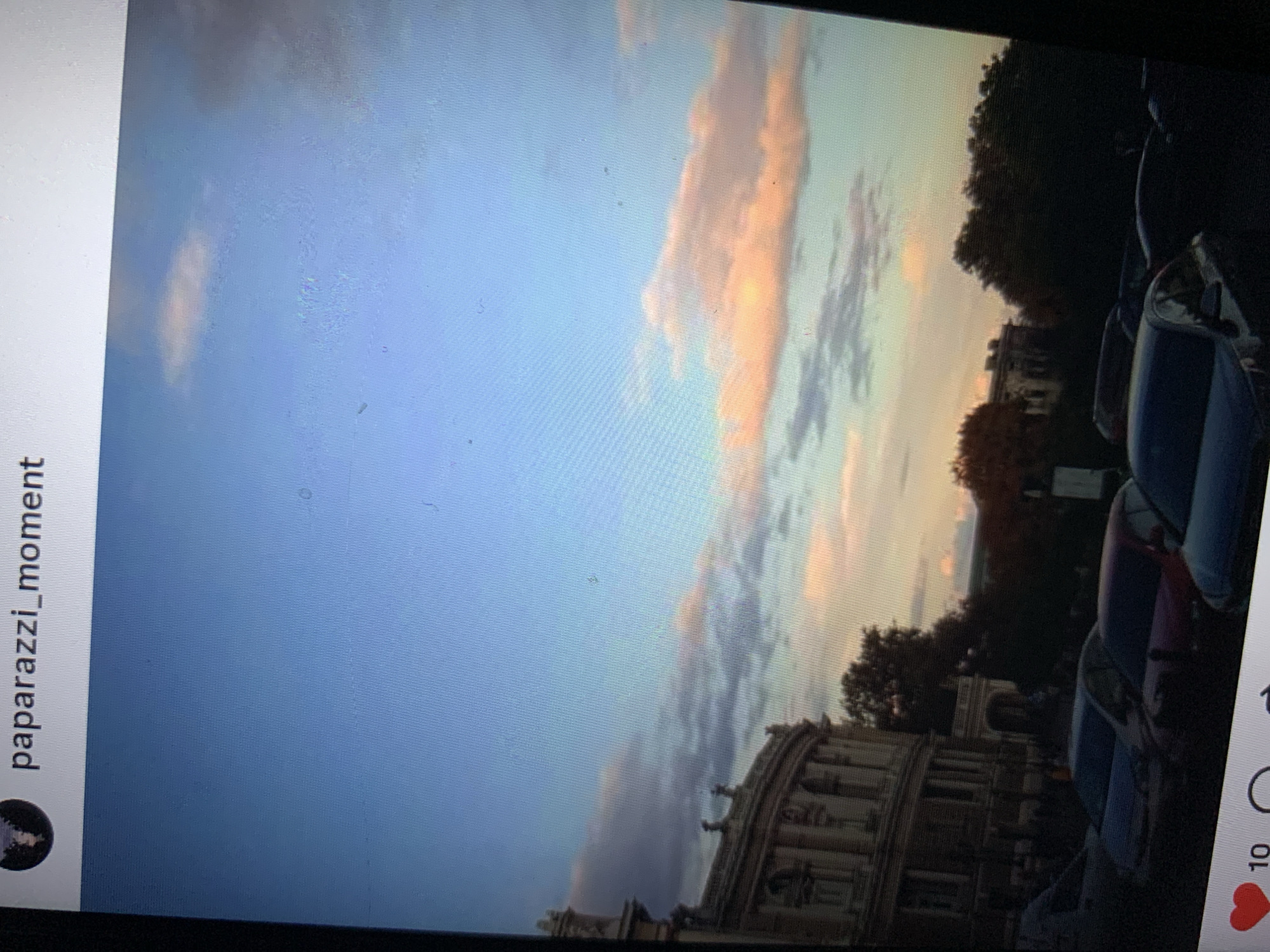 The image shows a wide shot of a street scene with a building on the left and parked cars in the foreground. The sky is a gradient of blue with pinkish clouds, suggesting it is either sunrise or sunset. The building appears to be historic with ornate architecture. Several cars are visible, parked along what looks like a street or parking lot. The environment is urban, with trees in the background. The Instagram username "paparazzi_moment" is displayed at the top of the image. The bottom of the image shows social media interaction icons: a heart, a number "10", and a circular icon. No people are clearly visible or identifiable. The scene is static.