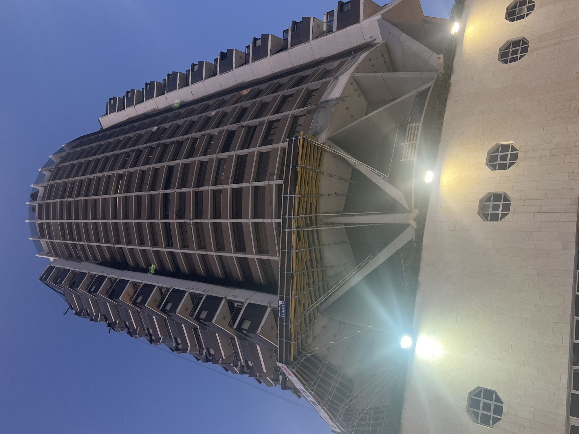 Here is a detailed description of the image:
The image depicts a tall, cylindrical building in Tel Aviv, Israel, constructed with a Brutalist architectural style. The building features a repeating pattern of rectangular windows that encircle its structure. The top of the building includes a smaller, rectangular section with protruding elements. Scaffolding is visible on one side of the building, indicating ongoing construction or maintenance.
Adjacent to the building is a shorter structure with a light-colored facade featuring a series of octagonal windows. The sky is a clear, deep blue, suggesting it may be either late evening or early morning. The angle of the image indicates that the photograph was taken from a low vantage point. The area appears to be lit by artificial light sources.