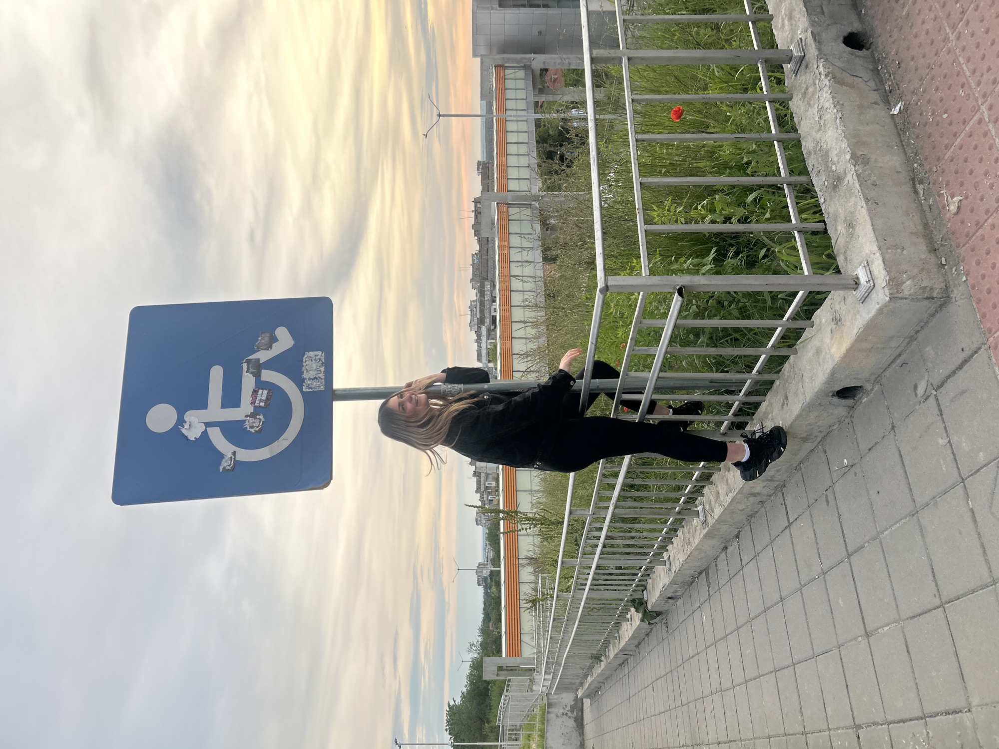 The image depicts a young woman posing on a metal railing next to a blue handicapped parking sign. The background shows a cityscape and a colorful sunset sky. The location appears to be near the city of Ruse, Bulgaria, based on architectural style and the general environment. The woman is wearing all black clothing and sneakers. She is leaning against the railing, partially obscuring the view beyond, and smiling. A single red poppy is visible in the vegetation behind the railing. The sidewalk appears to be paved with gray and red rectangular tiles. The handicapped sign is attached to a metal pole. Stickers or markings are present on the sign's surface.