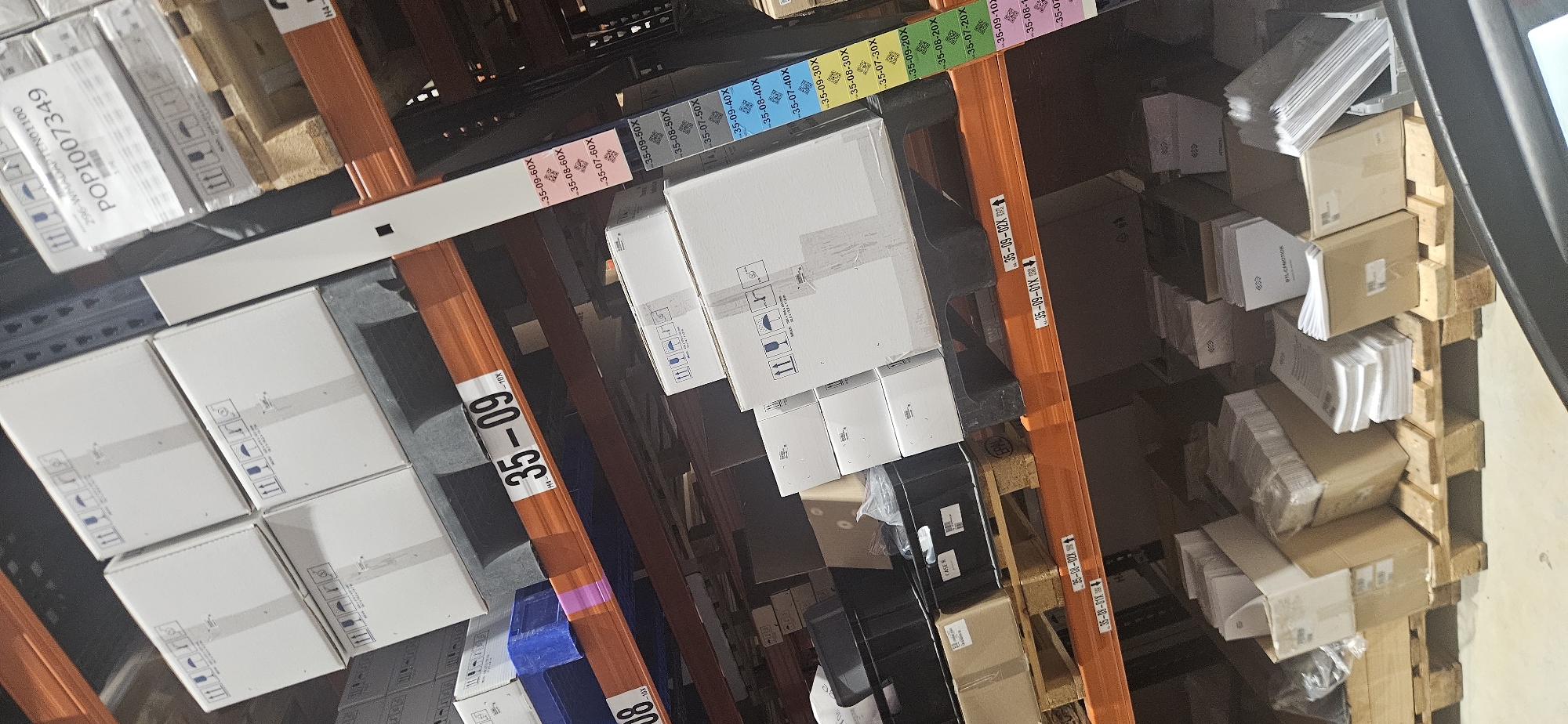 The image shows a view within a warehouse or storage facility, focusing on shelving units filled with various boxes and packages. The shelves are labeled with alphanumeric codes, such as "35-09-10X" and "35-08-01X." The items on the shelves vary in size and shape, including white rectangular boxes and smaller cardboard packages. The scene has industrial lighting, suggesting a practical, functional environment. The location appears to be an indoor storage area, though specific details indicating a location in Plovdiv, Bulgaria are absent.
