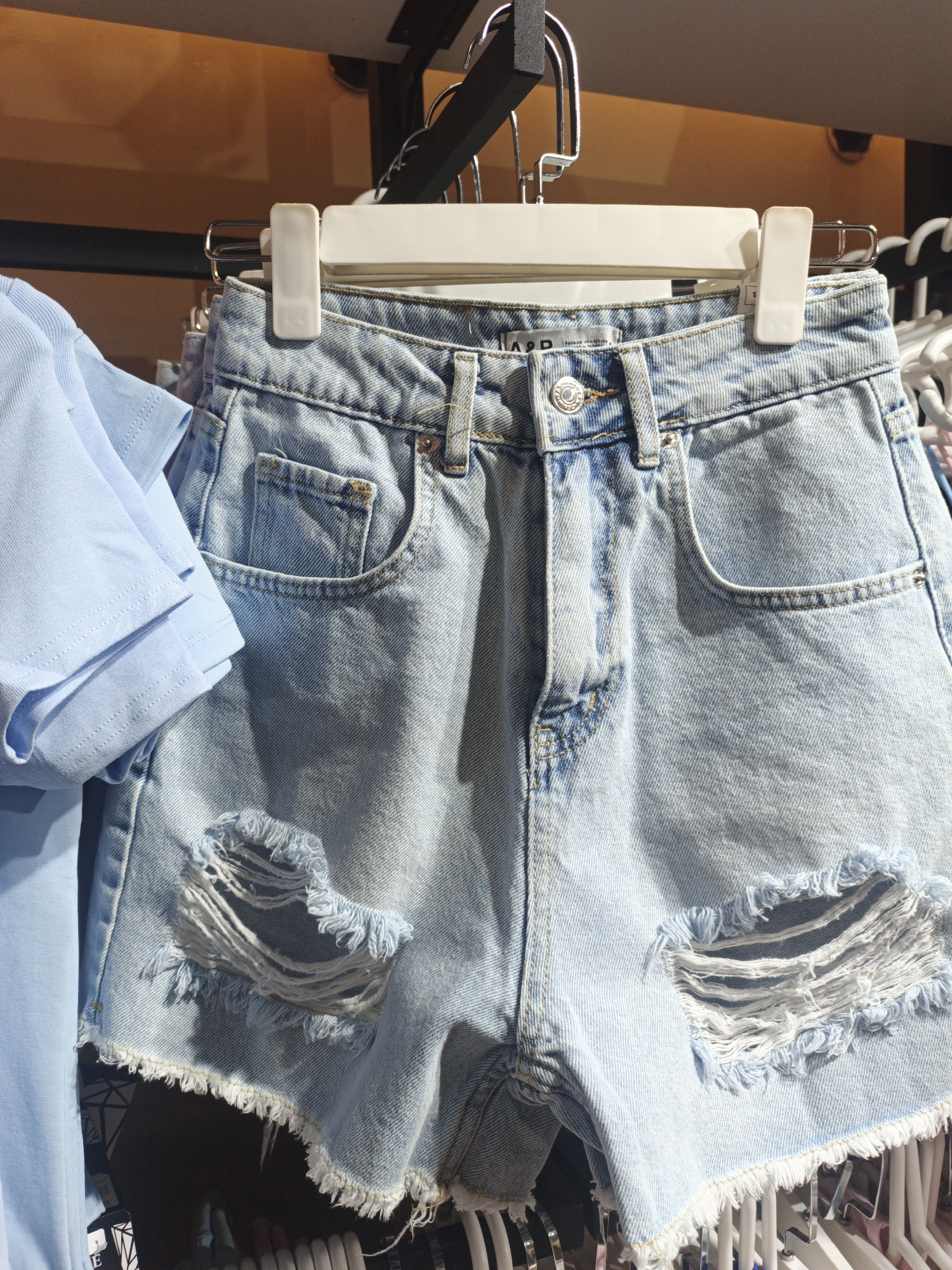 The image shows a close-up of light-wash denim shorts with rips and frayed edges, hanging on a white hanger. To the left of the shorts, a light blue t-shirt is also visible, folded and draped over its own hanger. The shorts appear to be displayed in a retail environment, possibly a clothing store. The background is blurred but suggests shelving and other clothing items. There are no people or specific identifying landmarks that would place this scene in Shymkent, Kazakhstan, or any other specific city. The scene is static and focused on the apparel.