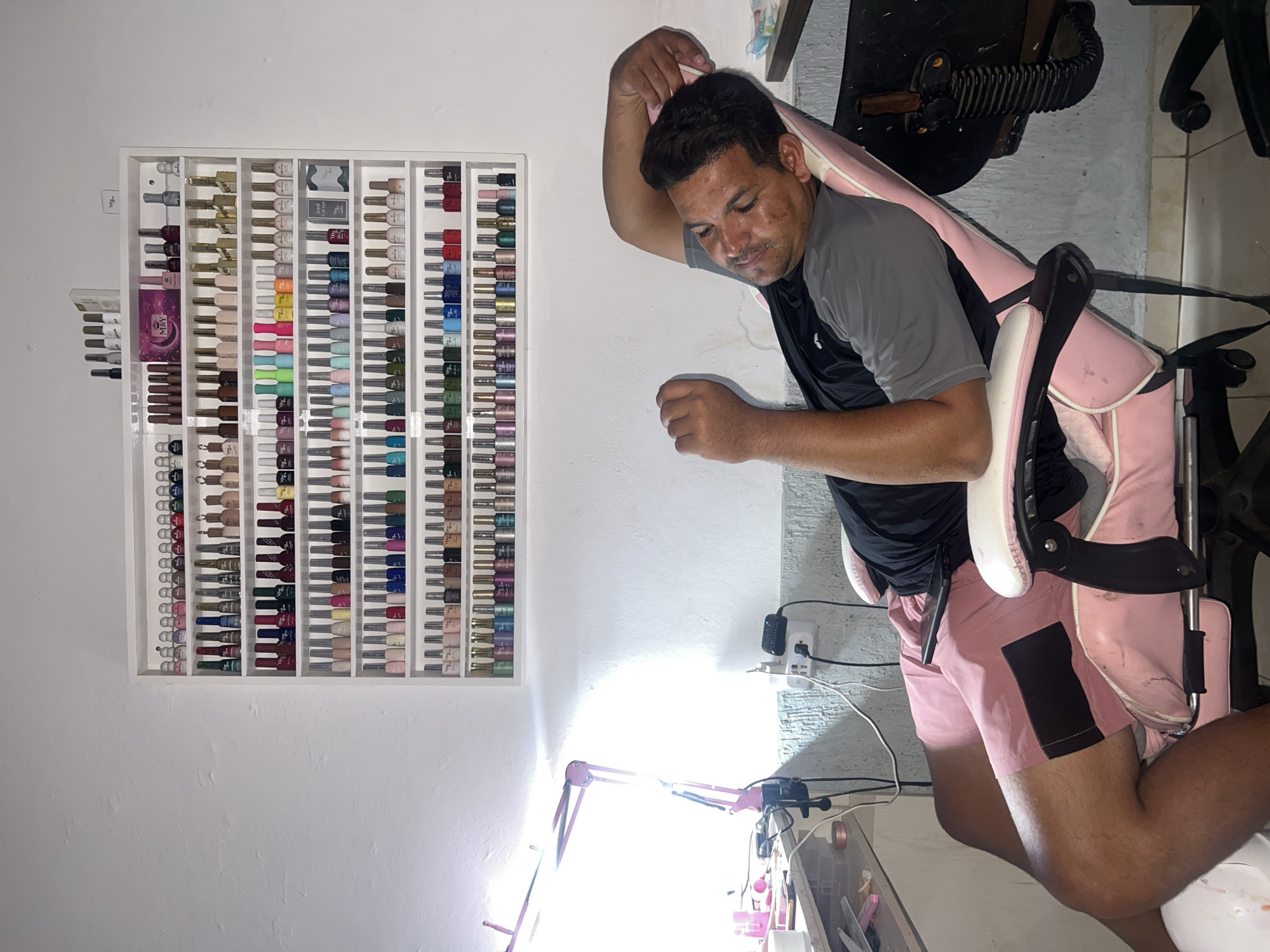 A man is reclined in a pink and white chair in what appears to be a nail salon in Juan Griego, Venezuela. He is wearing a gray t-shirt and pink shorts, with a phone resting on his lap. Behind him, a white shelf is mounted on the wall, filled with numerous bottles of nail polish. To the left, a pink adjustable desk lamp is illuminated, casting a bright light on a cluttered desk with various beauty supplies. Electrical outlets are visible on the wall behind the man's chair.