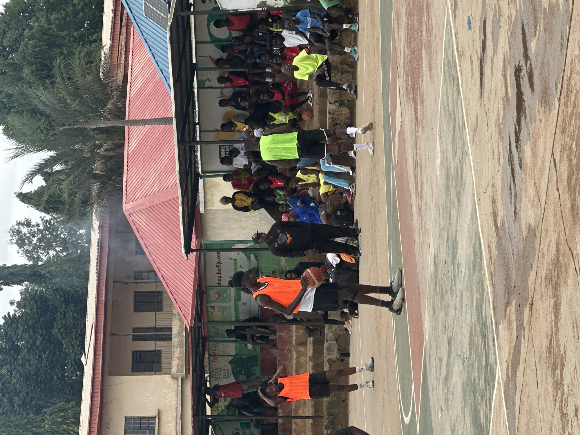 An outdoor basketball court in Jos, Nigeria, hosts a practice session. The court features worn concrete with painted lines and several puddles, indicating recent rain. Spectators are seated on concrete bleachers under a covered structure with red and blue corrugated metal roofs. The background shows a two-story building with a beige facade and balconies.
Several young men and women are on the court, many wearing orange or lime green bibs over athletic wear. One individual in an orange bib is holding a basketball, poised for a shot. Another person in a lime green bib faces away from the camera, also on the court. Spectators are dressed casually, some in team colors or bright athletic attire. The overall atmosphere suggests a spirited community event or training session.