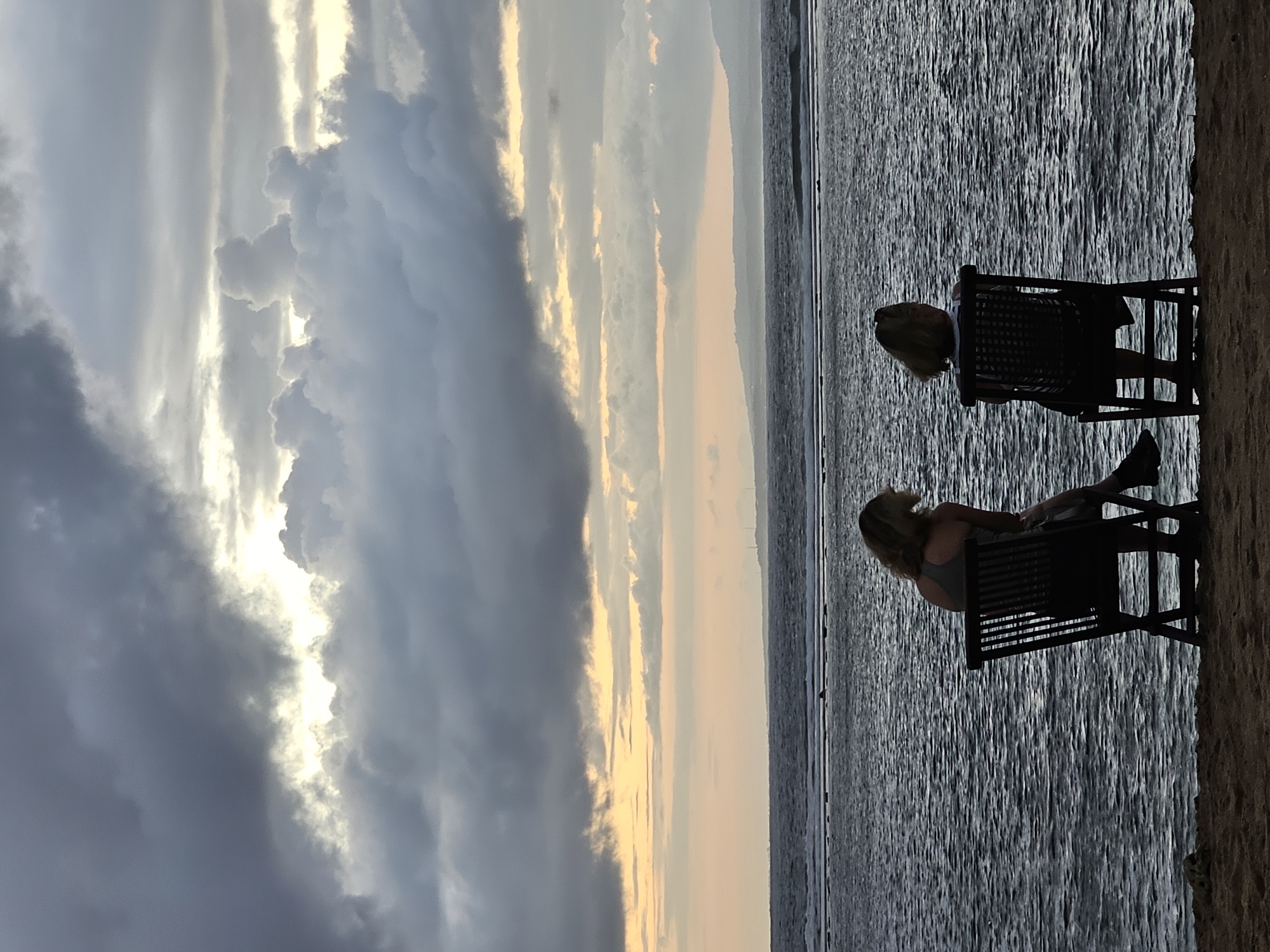 Two individuals are seated in chairs on a sandy beach, facing a body of water. The individual on the left is wearing a tank top and shorts, while the individual on the right is wearing a striped top and shorts. The sky above is filled with dramatic, dark clouds, with patches of light breaking through. The ocean appears choppy, with visible waves. In the distance, a silhouette of land with hills can be seen. The scene is dimly lit, suggesting it is either dawn or dusk. The image was captured at Denpasar, Indonesia.
