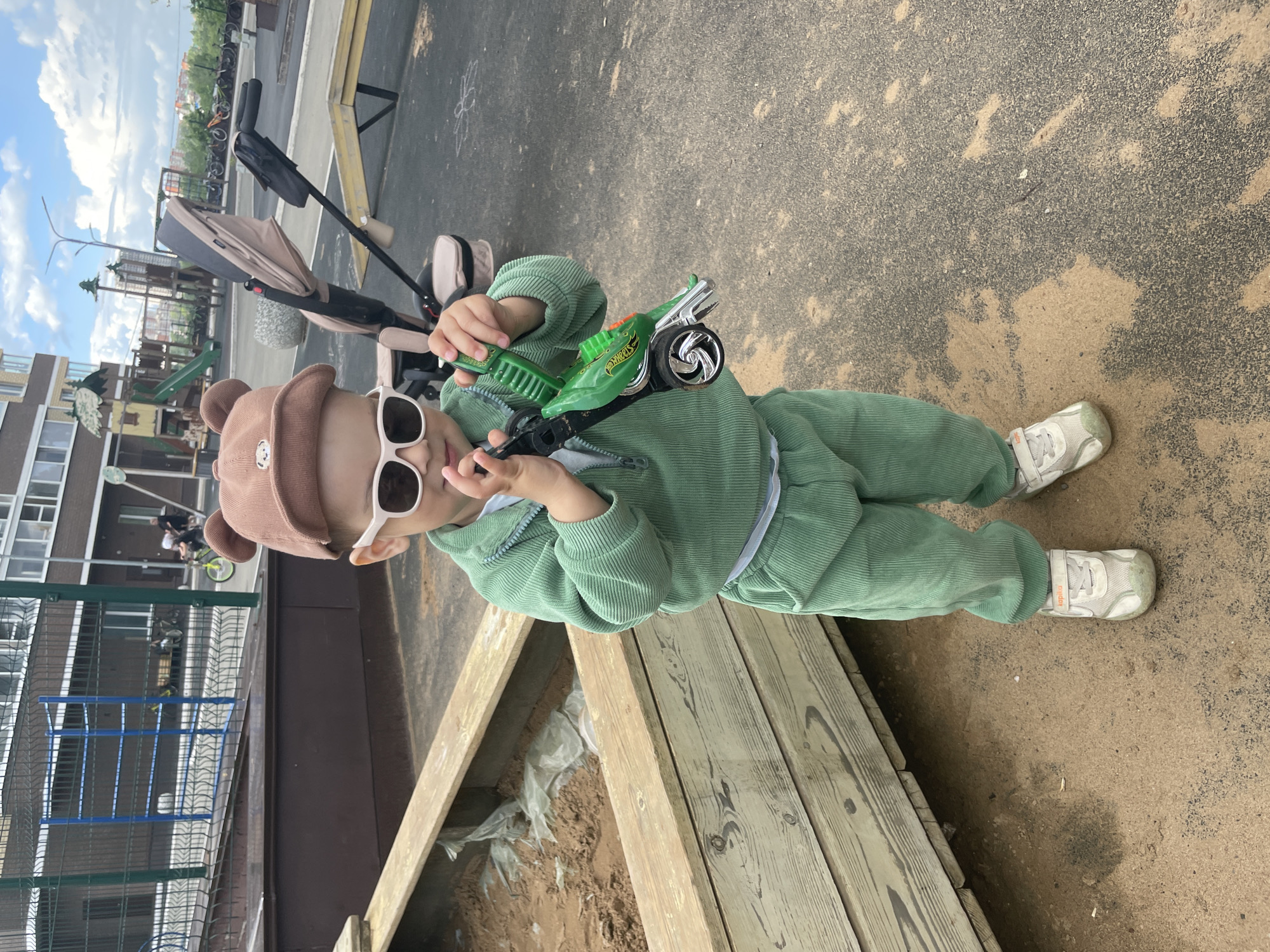 A toddler is standing in what appears to be a playground in Izhevsk, Russia. The child is wearing a green corduroy outfit, a brown bear hat, and white-rimmed sunglasses. They are holding a green toy car. A stroller is partially visible in the background. The playground setting includes wooden structures, a metal fence, and paved areas.