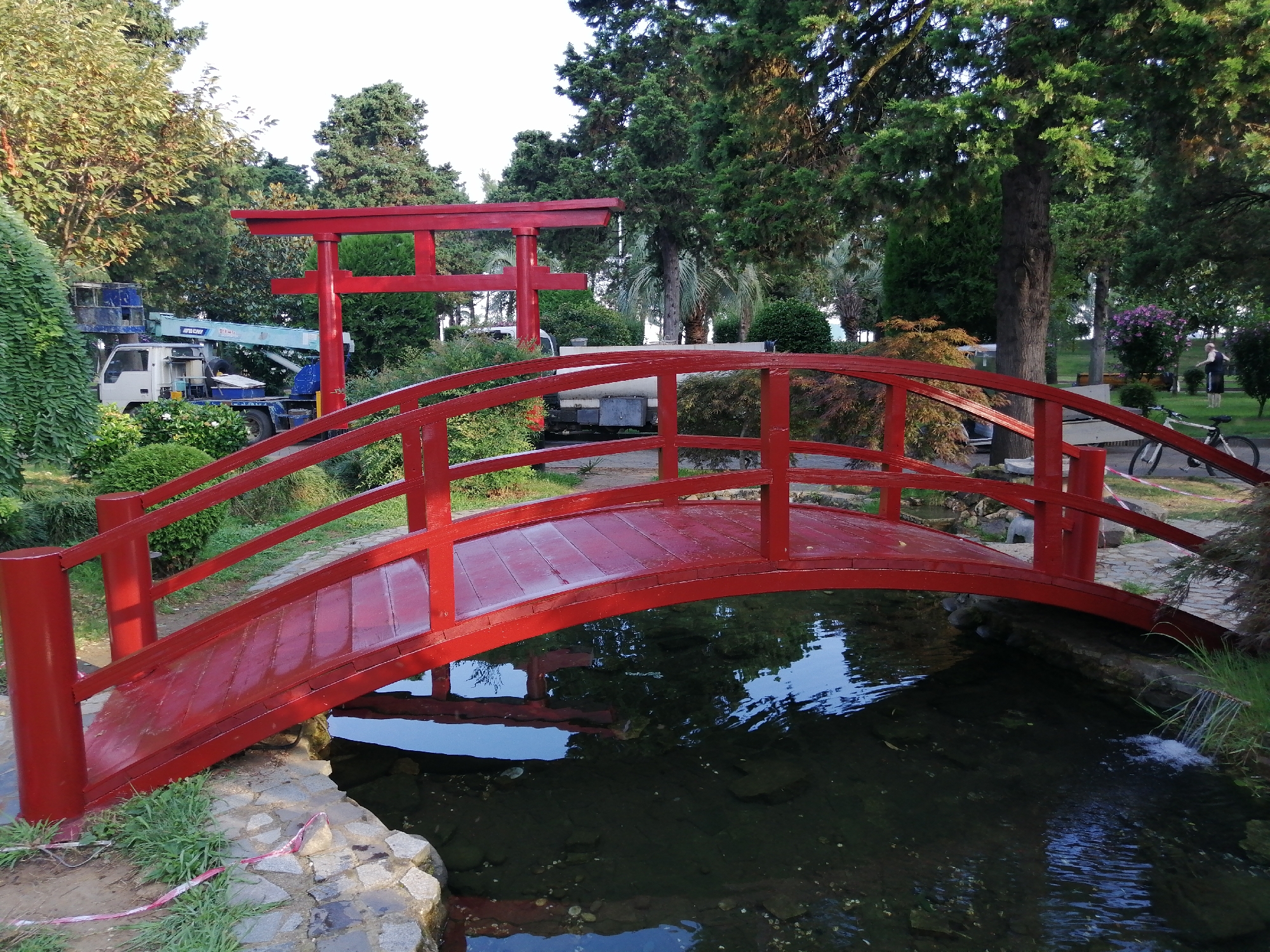 This image depicts a red wooden arched bridge over a small, dark water channel in Batumi, Georgia. Behind the bridge, a red torii gate stands prominently. To the left of the torii gate, a white utility truck with a blue lift mechanism is visible amongst lush green foliage. The background features a variety of trees, including evergreens and palm trees. In the distance, a person in dark clothing and a white bicycle are present near a cluster of purple flowers. The scene suggests a public park or garden setting.