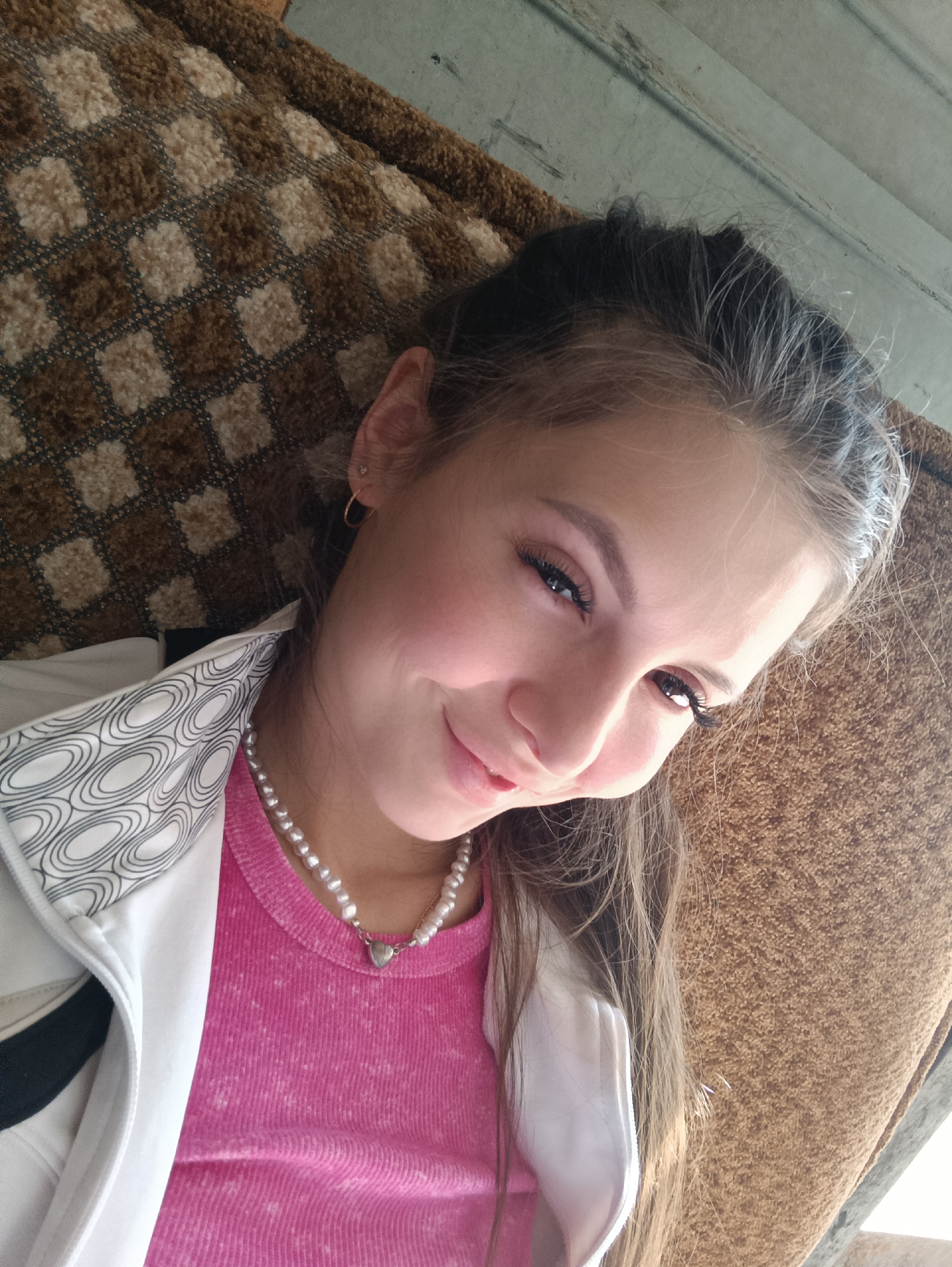 A young woman is lying on a brown patterned sofa indoors. She is wearing a pink shirt, a white jacket, and a pearl necklace with a heart pendant. She has dark hair pulled back and is smiling at the camera. The location appears to be in the city of Unknown, Unknown.