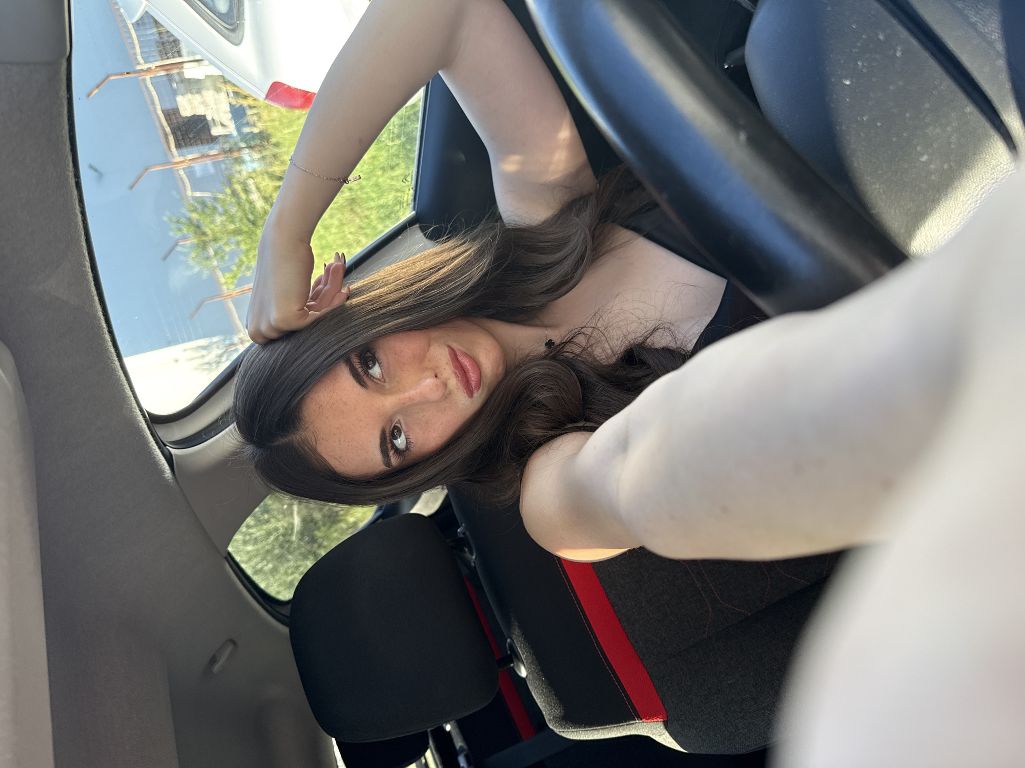 A young woman is taking a selfie inside a car. She has long, wavy brown hair, fair skin, and dark eyes. She is wearing a black top and a delicate bracelet on her left wrist. Her head is tilted, and she looks directly at the camera. The car's interior is visible, including the black steering wheel, black and grey car seats with red accents, and a rearview mirror. Outside the car window, buildings and trees are visible, suggesting an urban environment. The reflections in the car window and the limited scope of the background do not provide enough detail to identify the specific location in Plovdiv, Bulgaria.