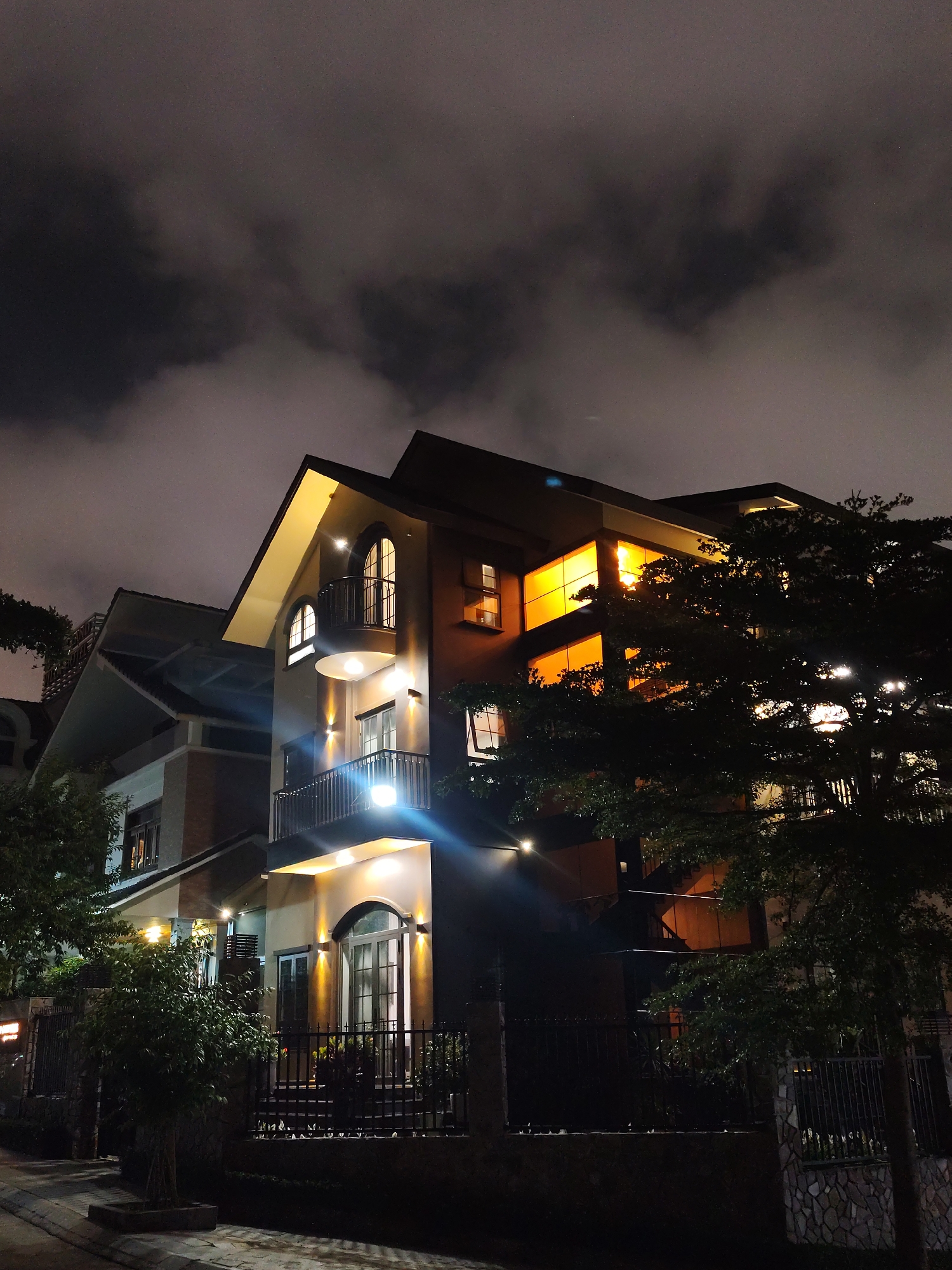 A nighttime exterior shot captures a modern, multi-story residence in Da Lat, Vietnam. The building features a light-colored facade on the lower levels and a darker brick or stained wood on the upper floors. Arched windows and balconies adorn the structure, illuminated by exterior lights. Warm orange light emanates from large windows on one side, suggesting interior illumination. A fence surrounds the property with foliage planted in front. Street lighting adds to the overall brightness of the scene. Above the building, clouds obscure much of the night sky.