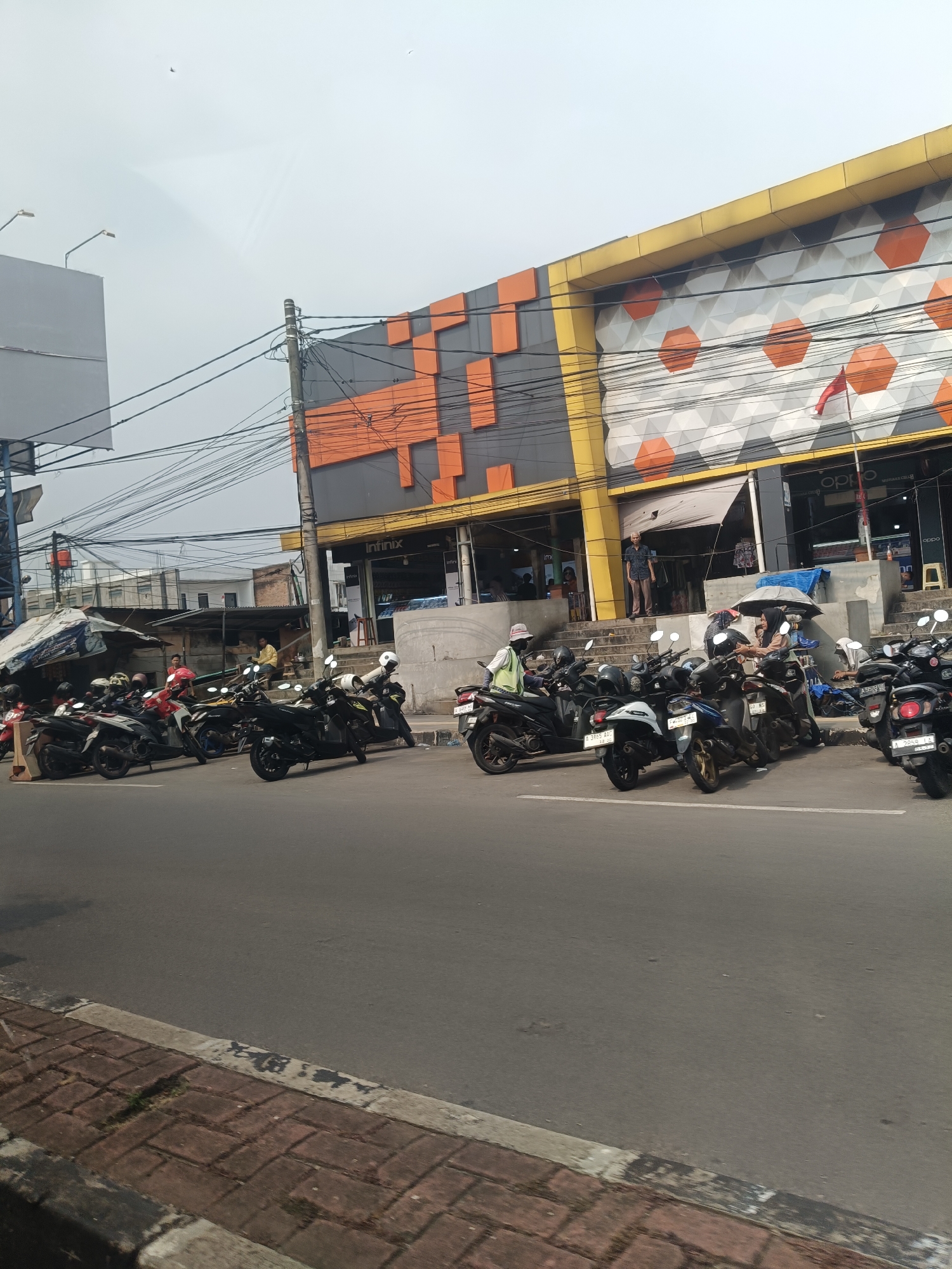 The image captures a street scene in Pandeglang, Indonesia, likely in front of a commercial establishment. A row of parked motorcycles lines the side of the road, suggesting a busy area with a need for vehicle parking. Behind the motorcycles, a building with a modern, geometric facade is visible, featuring orange and grey hexagonal patterns against a yellow trim. Signs for "Infinix" and "OPPO" are present, indicating the building likely houses electronics or mobile phone stores.
Several individuals are depicted in the scene. One person, wearing a light green vest and a white helmet, is seated on a motorcycle. Others are seen standing or sitting near the entrance of the building, some under an umbrella. The overall environment appears to be an urban setting with overhead power lines crisscrossing the sky. The road is paved, and a brick sidewalk is visible in the foreground. The weather appears overcast.