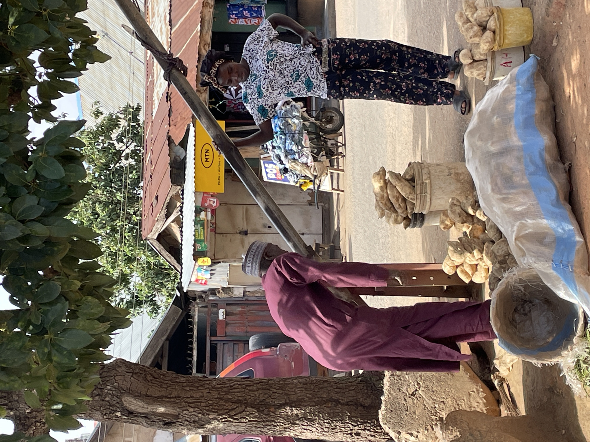 A street scene in Jos, Nigeria, depicts two individuals amidst a backdrop of local commerce and daily life. A woman wearing a patterned blouse and dark floral pants stands beside a metal pole attached to a yellow MTN sign, possibly engaging in a transaction or conversation. Buckets and a large sack filled with root vegetables, likely yams or sweet potatoes, sit nearby, suggesting she is selling them. A man in a maroon robe and cap gestures with his arm, possibly interacting with the woman. The environment includes a mix of corrugated metal structures, vehicles, and trees, indicative of a bustling roadside marketplace in Jos.