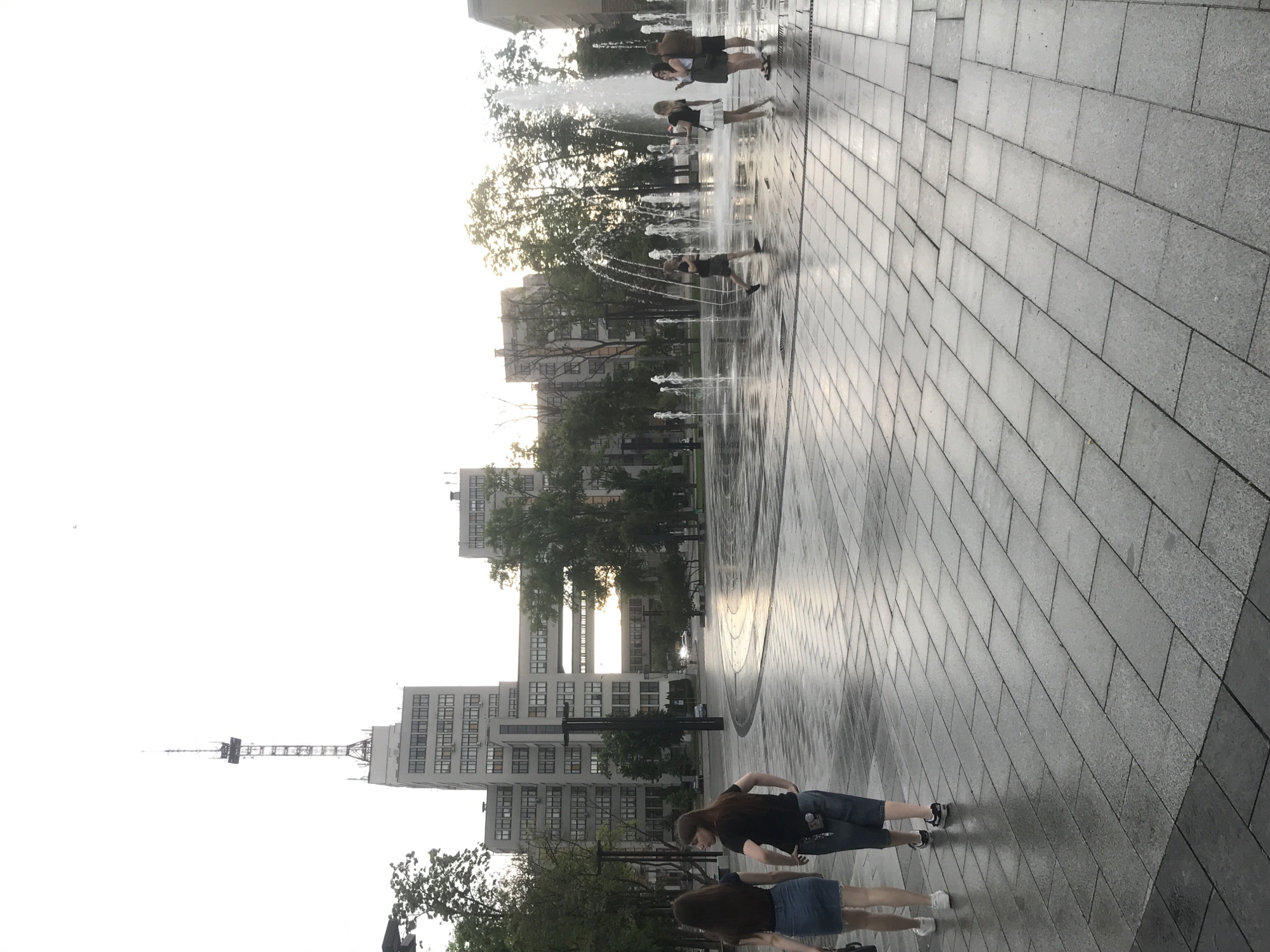 The image shows a street scene in Kharkiv, Ukraine, with a fountain feature on a gray stone-tiled surface. Several individuals are visible, some interacting with the fountain jets of water, others walking along the tiled area. In the background are apartment buildings and trees, and a tall tower with a metal framework is also visible. The sky is overcast, suggesting it is either early morning or late afternoon.