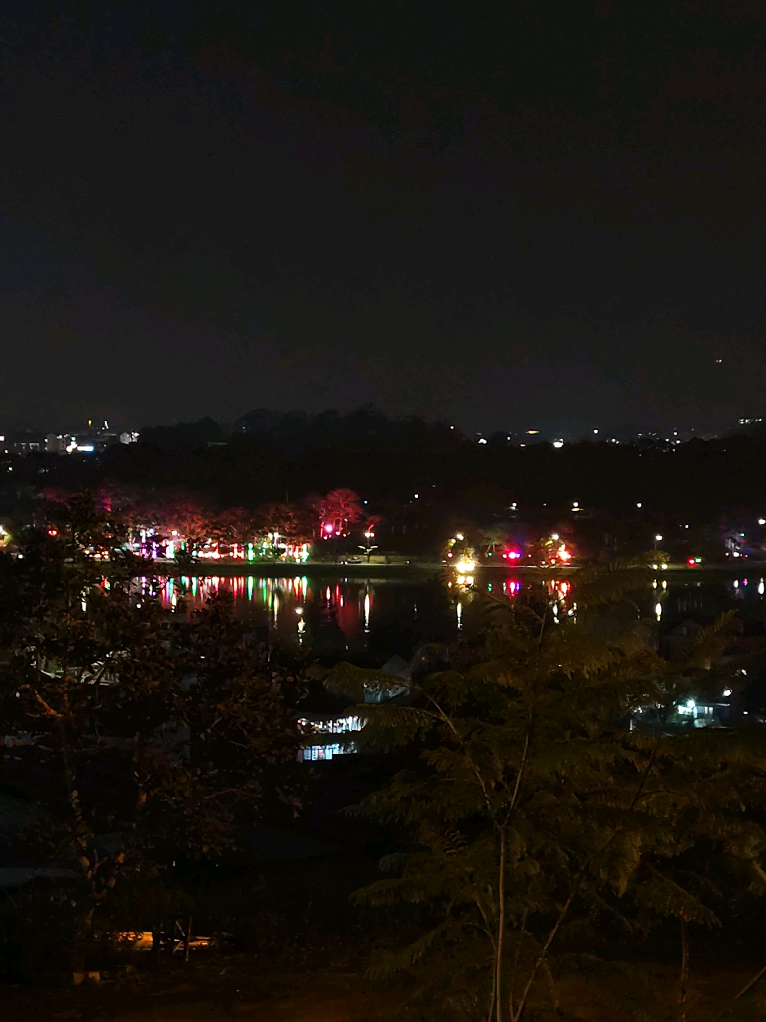 A nighttime aerial view captures a tranquil lake in Da Lat, Vietnam, lined with trees illuminated by colorful festive lights. The reflections of the lights shimmer on the water's surface, creating a vibrant spectacle against the dark sky. In the background, distant city lights dot the horizon, hinting at the presence of more urban development beyond the immediate lakeside area. The foreground is dominated by the dark silhouettes of trees, adding depth and a sense of seclusion to the scene. No people are visible in the image.