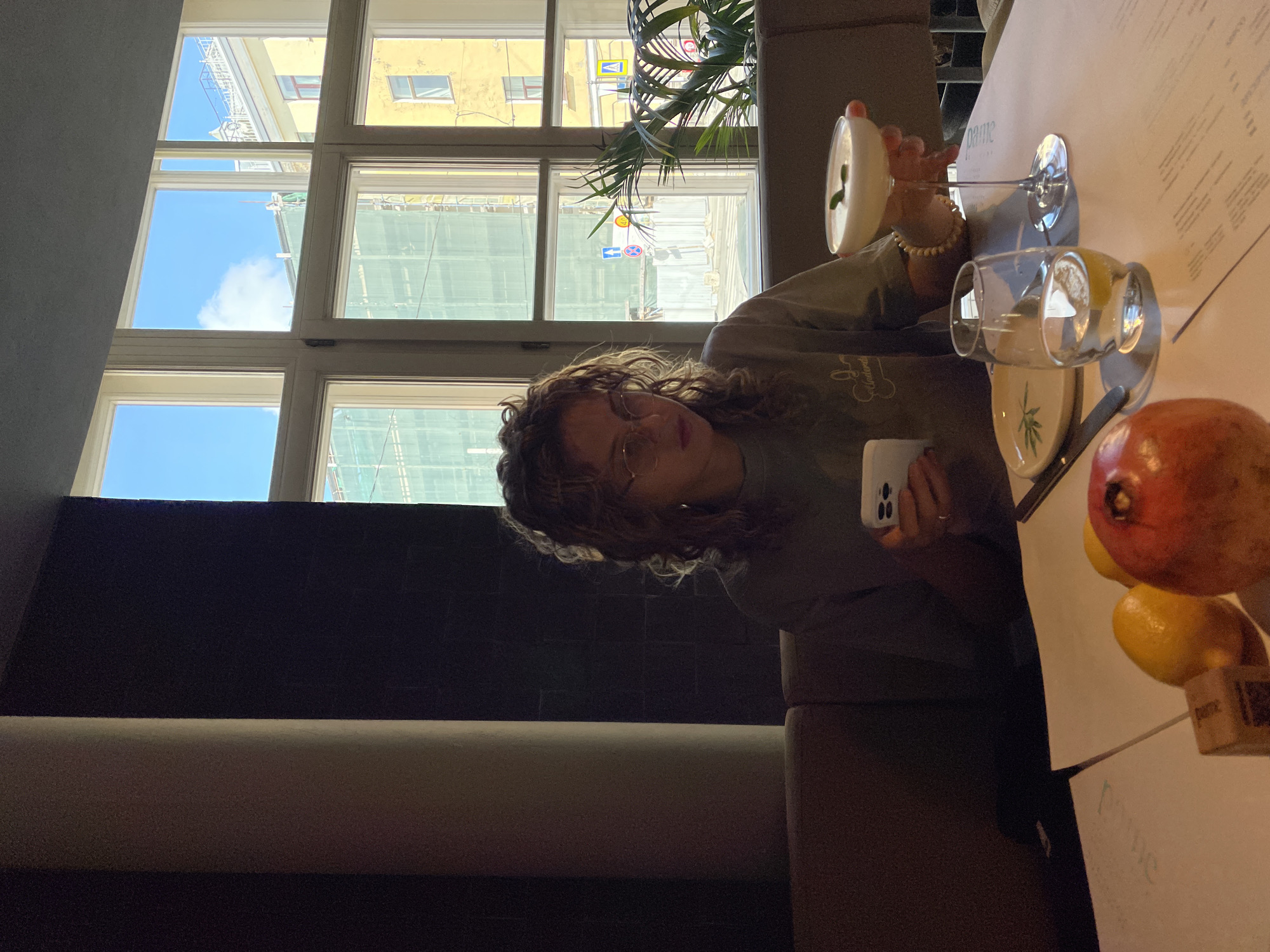 A woman sits at a table next to a window in a building in Saint Petersburg, Russia. The window offers a view of neighboring buildings, one of which is under construction or renovation, with scaffolding visible. The sky is partly cloudy. The woman, with curly hair and glasses, is holding a white smartphone in her left hand. On the table, there are various items: a cocktail glass with a drink, a glass of water, a plate, a knife, a pomegranate, lemons, and a menu with the word "pame" visible on it. The environment is indoor, likely a restaurant or cafe, with neutral color tones. The table is situated next to a dark-colored wall with horizontal beige stripes. A potted plant is visible near the window.