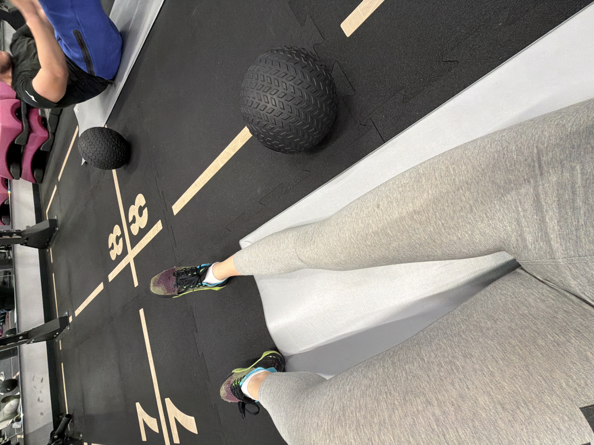 The image shows an indoor workout area. There are two people present. One person is lying on a mat in the upper left, wearing a black shirt and blue pants. The other person, closer to the camera, is wearing light gray leggings and athletic shoes with colorful accents. A gray, cushioned bench is between the two legs. Two textured black medicine balls are on the floor, one larger than the other. The floor is covered in black gym mats with white markings. The markings include numbers and lines. To the left of the frame is a weight rack with various attachments. The location is possibly a gym. The photo was taken indoors. It is impossible to determine if the image was taken in the city of Pernik, Bulgaria.