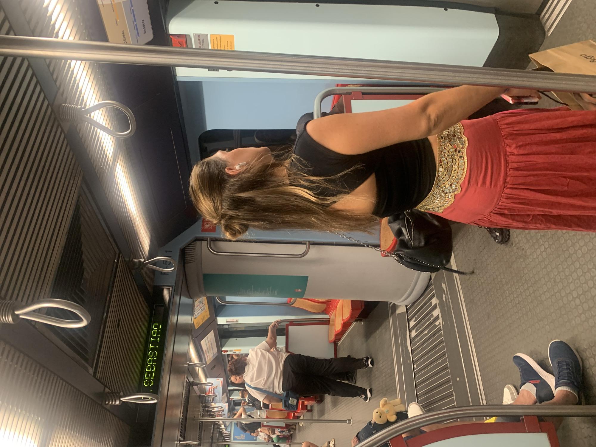 The image shows the interior of a subway car, likely on the Lisbon Metro, given the visible signage. In the foreground, a woman with her back to the camera is wearing a black top and a long, flowing red skirt with a bejeweled belt. She has her hair styled in a bun and is wearing white earbuds. To her left, another woman in a white shirt and dark pants stands, holding a blue bag. In the background, other passengers are visible, some seated and others standing. A yellow teddy bear toy is placed on the floor in the middle ground. The subway car features red and orange seats, silver grab handles on the ceiling, and a digital display showing "SEBASTIÃO" in green text. The floor is gray and textured. The overall environment suggests a public transportation setting within the city of Lisbon.