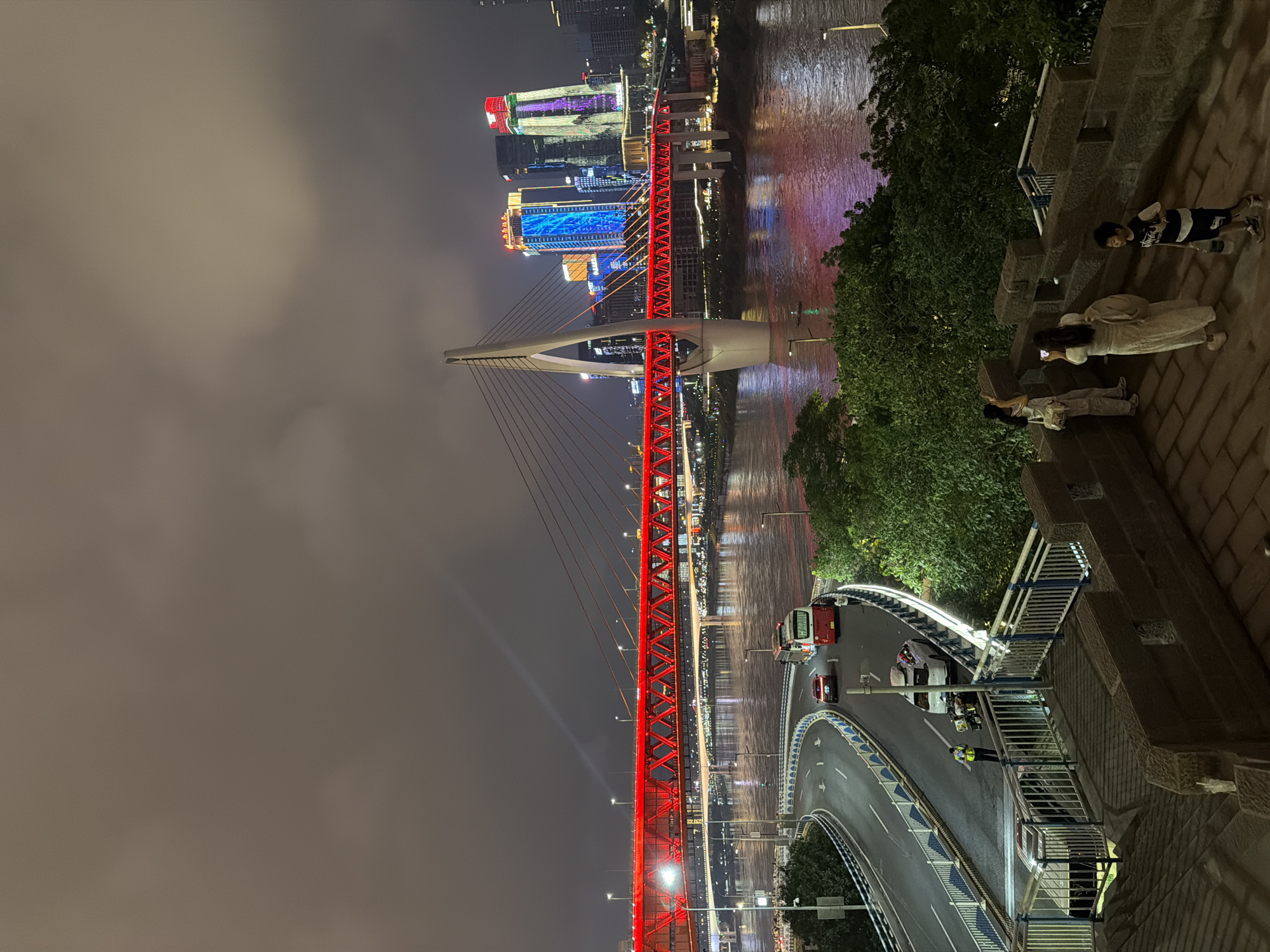 The image depicts a nighttime cityscape in Yuzhong District, China, dominated by a brightly lit red suspension bridge spanning a wide river. The bridge is a central element, with its illuminated structure and suspension cables creating a vibrant spectacle against the dark sky. In the background, modern skyscrapers are visible, adorned with colorful, dynamic light displays, contributing to the urban glow.
In the foreground, a stone wall and steps suggest a vantage point overlooking the scene. On this elevated area, three individuals are present. A woman in a light-colored dress and a backpack is standing, holding a smartphone and appearing to take a photograph. Another person, likely a woman, stands beside her, also holding a phone and facing the bridge. A young boy in a dark t-shirt and shorts is walking away from the camera, holding a bottle.
Below the vantage point, a curved road is visible, carrying vehicular traffic. A white car is stopped on the road, and a police officer in a yellow vest is standing nearby, interacting with individuals. A red bus is also on the road, with several passengers visible through its windows. The river reflects the city lights, creating a shimmering surface. Lush green trees are interspersed throughout the scene, softening the urban landscape. The overall atmosphere is one of nighttime urban activity and illuminated architecture.