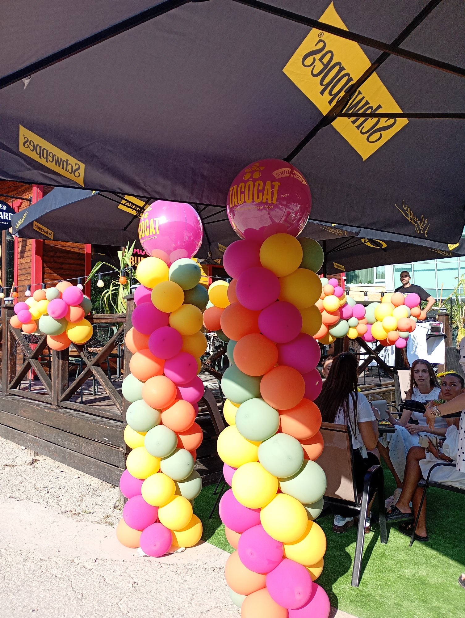 The image depicts an outdoor seating area with a wooden deck and railing, set up for an event. Two tall balloon columns, adorned with pink, yellow, orange, and light green balloons, stand prominently in the foreground. One column is topped with a large pink inflatable sphere featuring the "TACOCAT" logo. In the background, several black umbrellas provide shade over tables where people are seated. A DJ booth is visible with a man standing behind it. The environment appears to be a public space, possibly a restaurant or bar, in Varna, Bulgaria, as indicated by the presence of the "Schwabs" logo on the umbrellas, a known establishment in Varna. The scene is brightly lit, suggesting it is daytime, and the overall atmosphere is festive and casual.