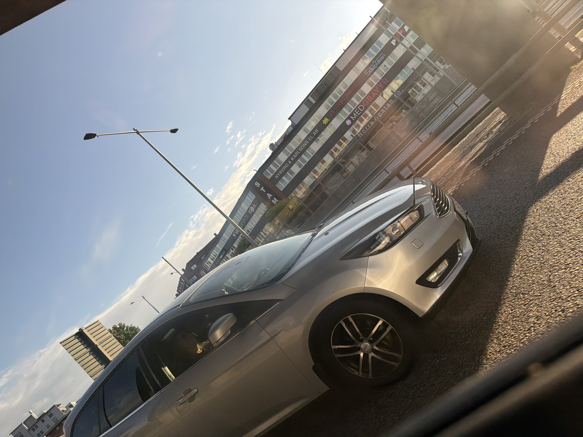 The image shows a silver Ford Focus parked on a street in Gothenburg, Sweden, with a building in the background. The building has signs for businesses such as "NYBERG & KARLSONS EL. AB," "MEDGRAVYR," and "Stadshusen." A street lamp is visible in the sky. The sun is low in the sky, casting long shadows, suggesting it is either early morning or late afternoon. The car appears to be in good condition. There are no people clearly visible in the car or its immediate vicinity. The street is paved with asphalt.