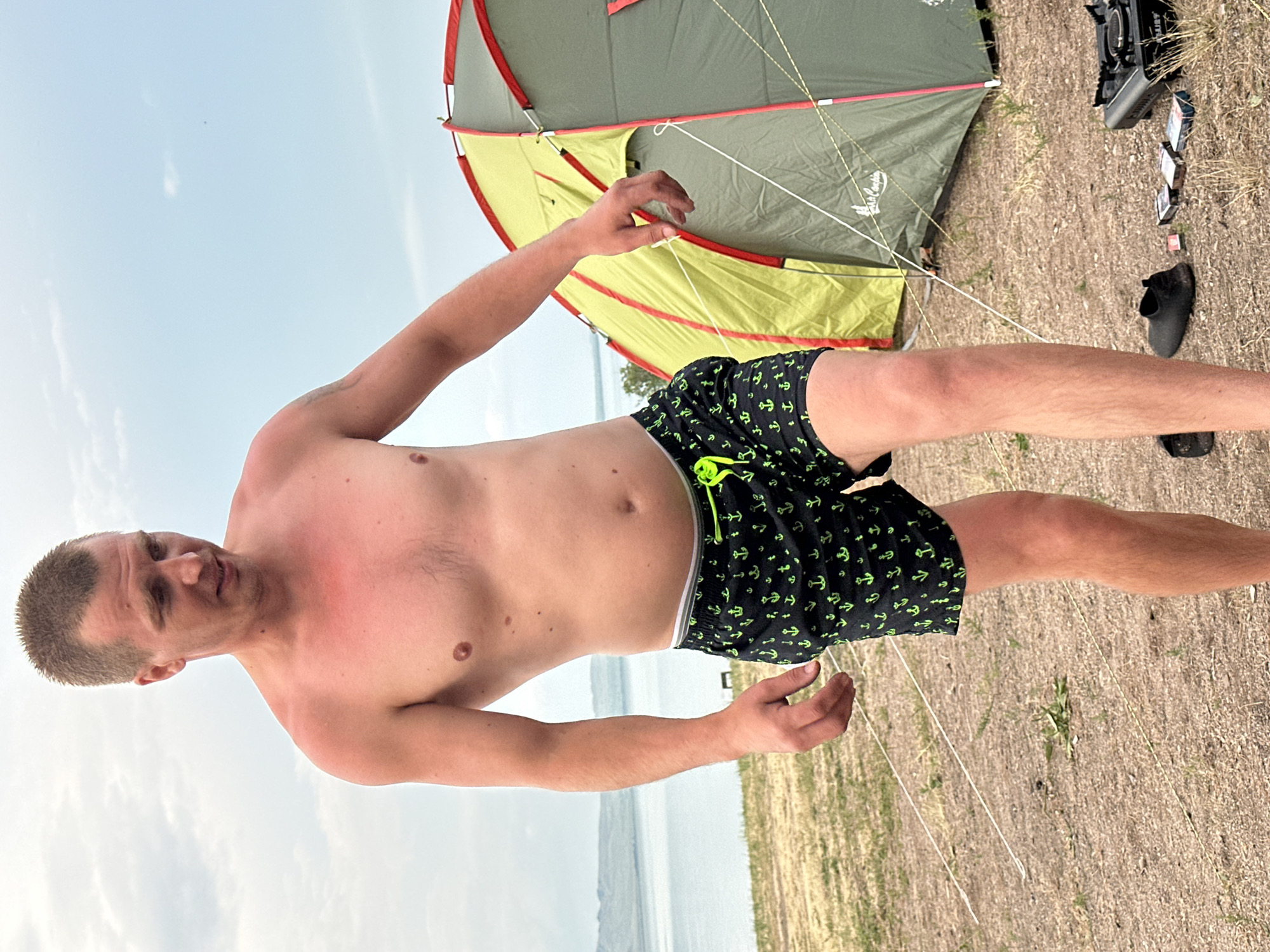 In an outdoor setting, a shirtless man wearing black swim trunks with a green anchor pattern stands on dry, sandy ground. He has a short haircut and is looking slightly off-camera with an impassive expression. Redness is visible on his upper chest and shoulders, suggesting sunburn.
Behind him, a partially set-up tent with green and yellow fabric is visible. The tent is secured by ropes. In the background, a body of water and a hazy sky can be seen. The overall scene appears to be a campsite near a body of water, likely during daylight hours given the bright sky. There is no visible text in the image.
