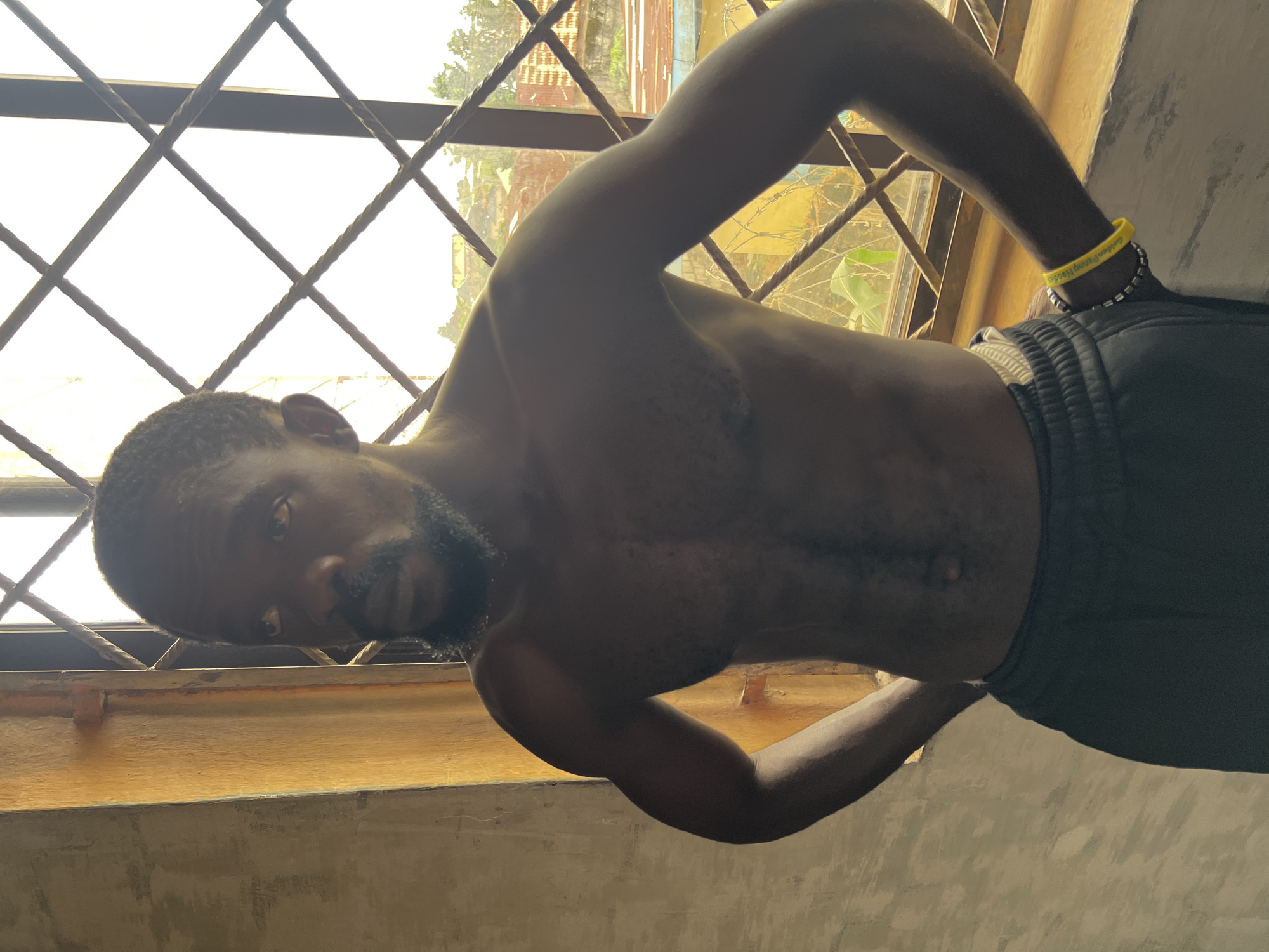 A shirtless Black man with a beard stands with his hands on his hips, facing a window with a diamond-patterned grate. The man is wearing dark sweatpants. A yellow and silver bracelet is visible on his right wrist. The window overlooks a distant cityscape with buildings and trees, bathed in sunlight. The man's left is a textured, beige wall. The scene appears to be indoors, possibly in a residence, in Jos, Nigeria.