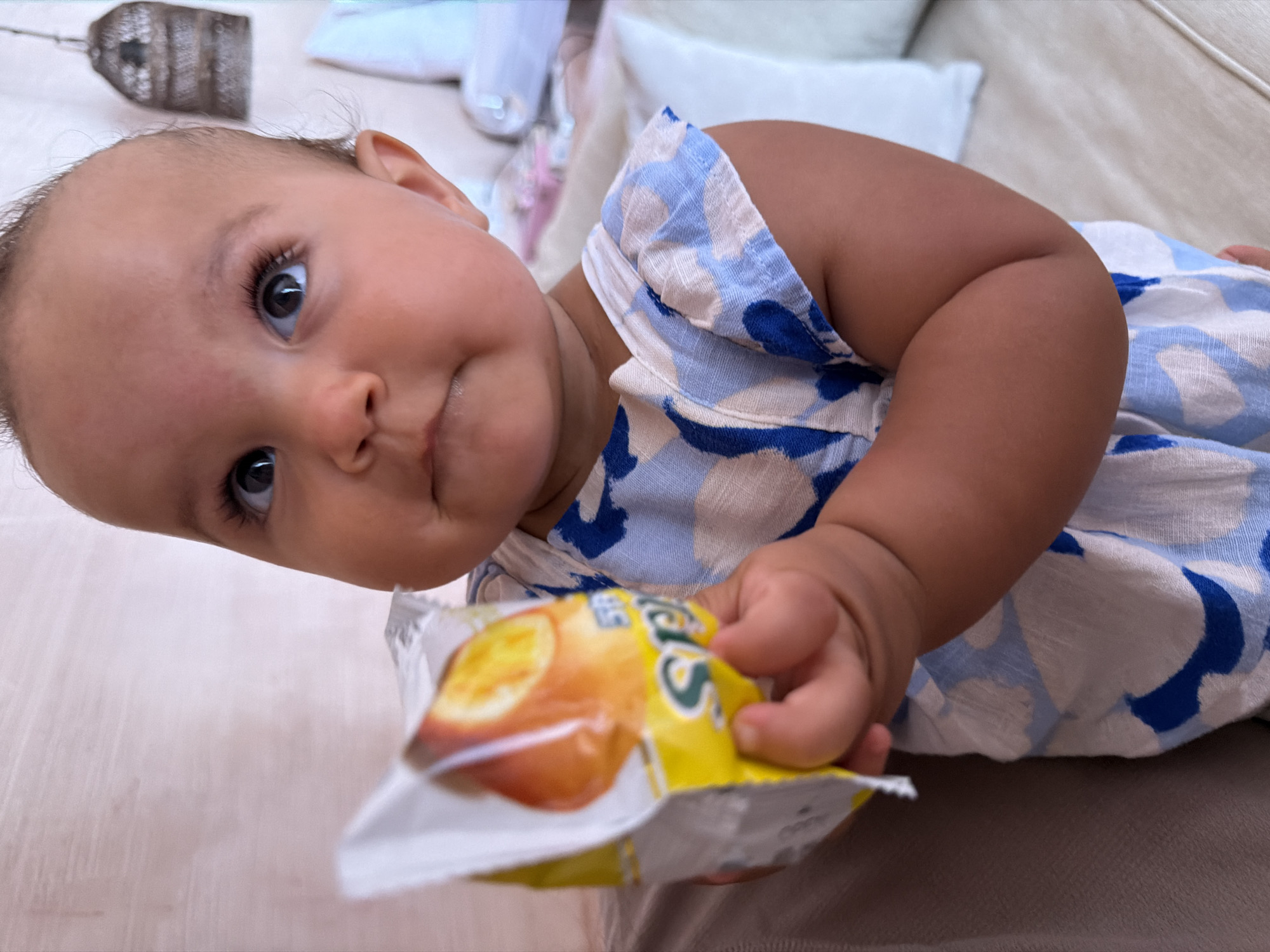 A baby lies on a beige surface, holding an opened, foil snack packet with a yellow and orange label. The baby wears a blue and white patterned dress. A decorative lantern and white pillows are visible in the blurred background. The photo was taken in North Kuta, Indonesia.