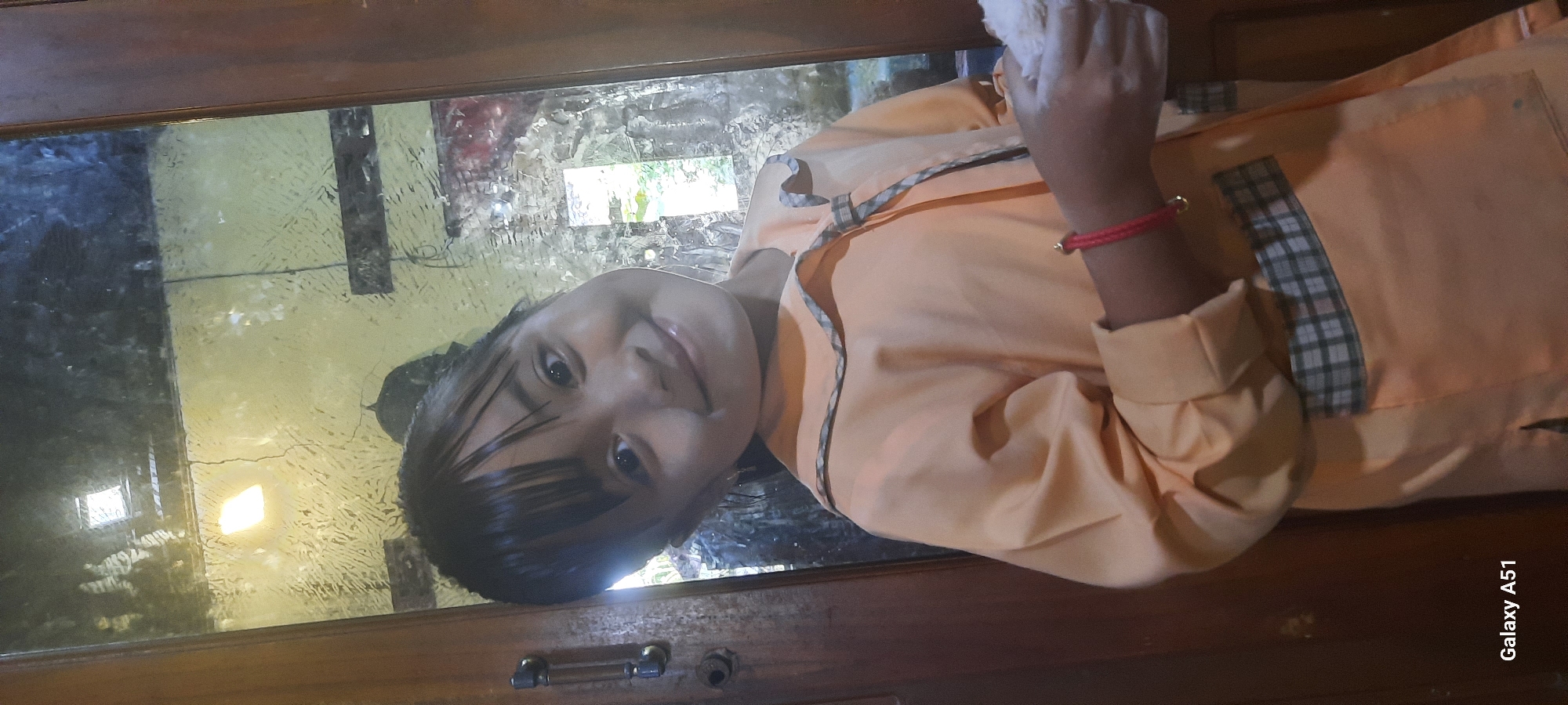The image shows a young girl, appearing to be of Asian descent, standing in front of a wooden door with a glass panel. She is wearing a light orange collared shirt with plaid trim on the collar and pockets. She has dark hair and is smiling at the camera. A red bracelet with a gold charm is visible on her right wrist. Her right hand is holding a white object, possibly fabric or paper, and appears to have white powder on it. The background, visible through the glass, is blurred and shows a light source, possibly a lamp, and some foliage. The text "Galaxy A51" is visible in the bottom left corner of the image, indicating the device used to take the photo. The location in the city of Jatitengah, Indonesia, is mentioned, though it is not visually evident from the image itself.