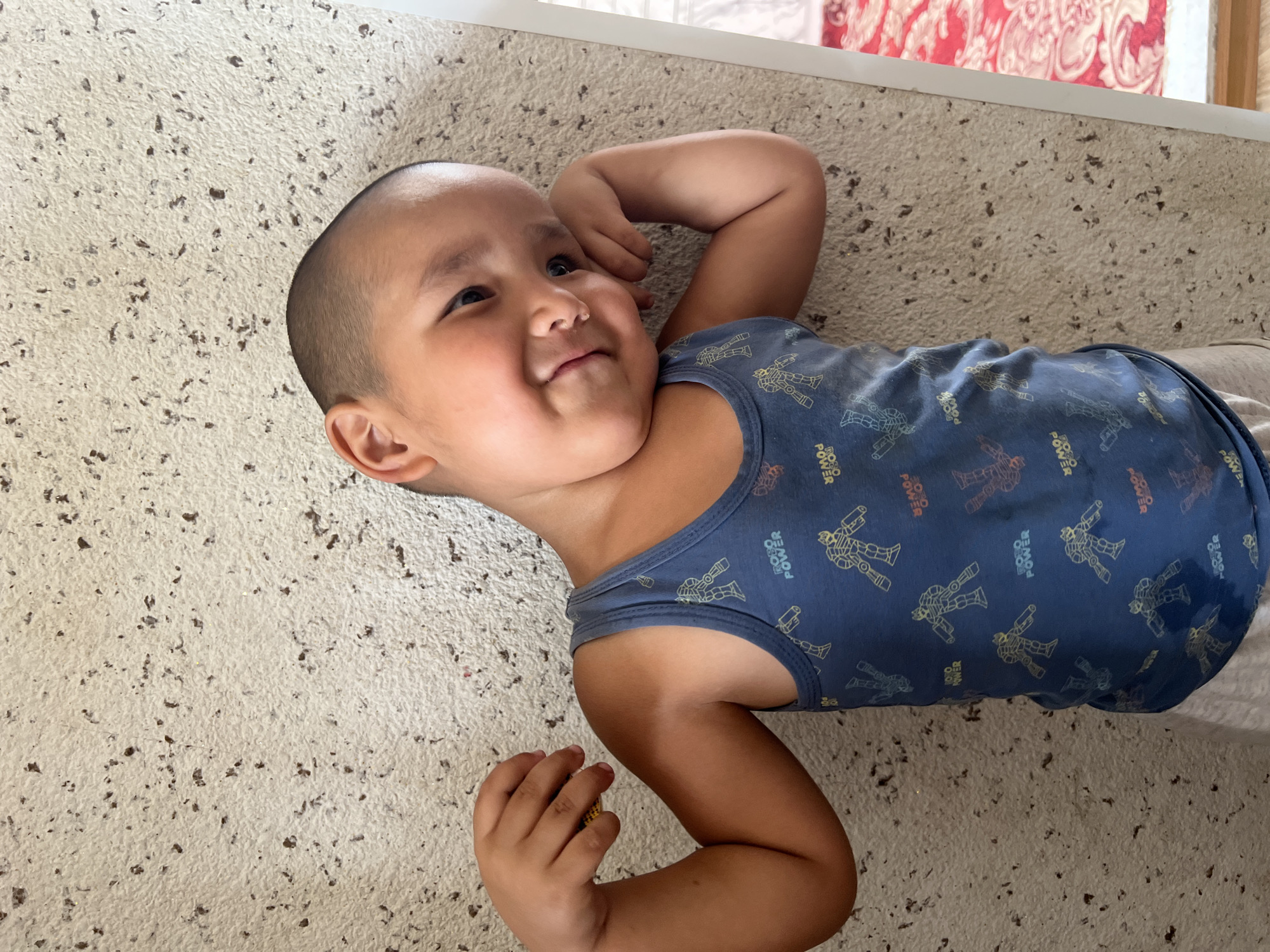 A young boy with a shaved head is leaning against a textured wall in what appears to be a home in Kara-Dıykan, Kyrgyzstan. He is wearing a blue sleeveless shirt with a pattern of robots and the words "ROBO POWER" printed on it, and light-colored shorts. His left arm is bent, with his hand resting near his head, and his right arm is bent at the elbow, with his fist near his bicep. He is smiling and looking towards the camera. The wall behind him is light-colored with dark speckles. To the right of the boy, a sliver of a red patterned rug is visible.