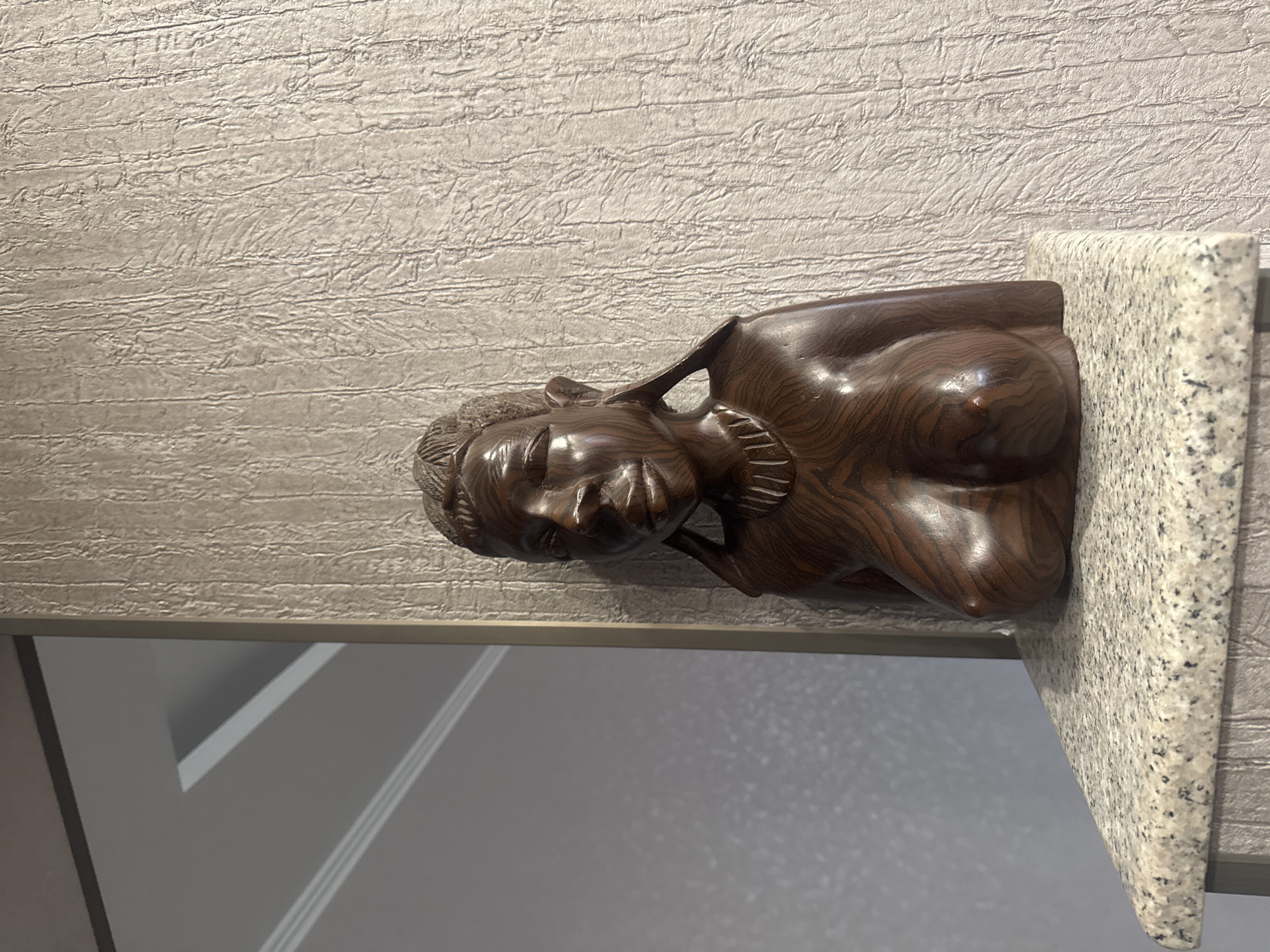 A dark brown wood sculpture of a reclining female figure sits on a light granite surface. The sculpture is resting on a ledge beneath a wall covered in textured, horizontal striped wallpaper. The wall and sculpture are angled to the left, revealing a glimpse of a gray wall with white trim in the lower left corner. The scene is devoid of any discernible identifying features that would suggest the location is in Tbilisi, Georgia.