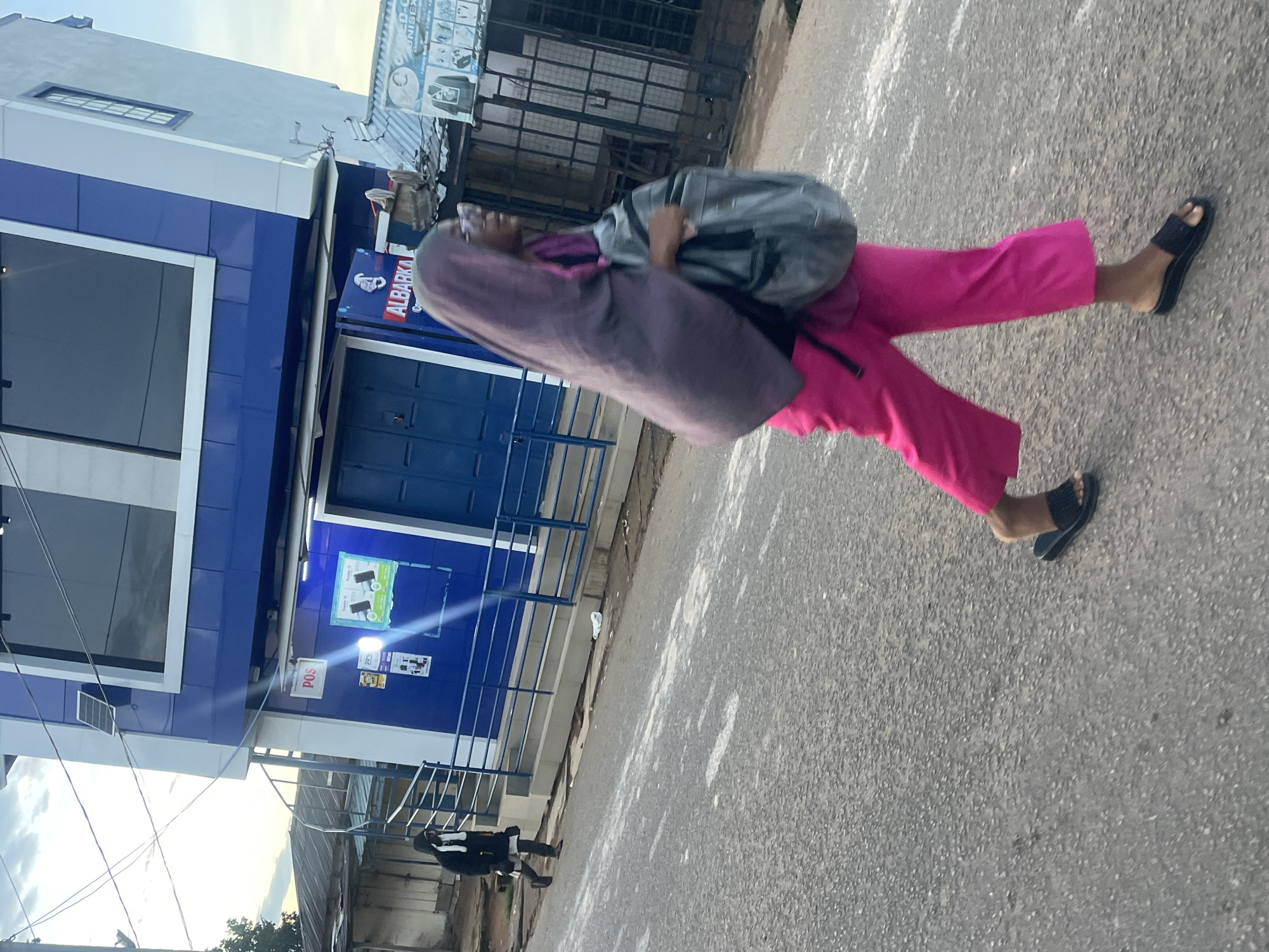 In Jos, Nigeria, on a paved street, a woman wearing a purple hijab and bright pink cropped pants walks from left to right across the frame. She is carrying a gray backpack on her right shoulder and wearing black slide sandals. In the background, a two-story blue and white building displays signs for "POS" and "ALBARKA". To the left of the building, another person in dark clothing walks away from the camera. The street is mostly empty, with some debris visible on the asphalt. The sky is overcast with thin clouds.
