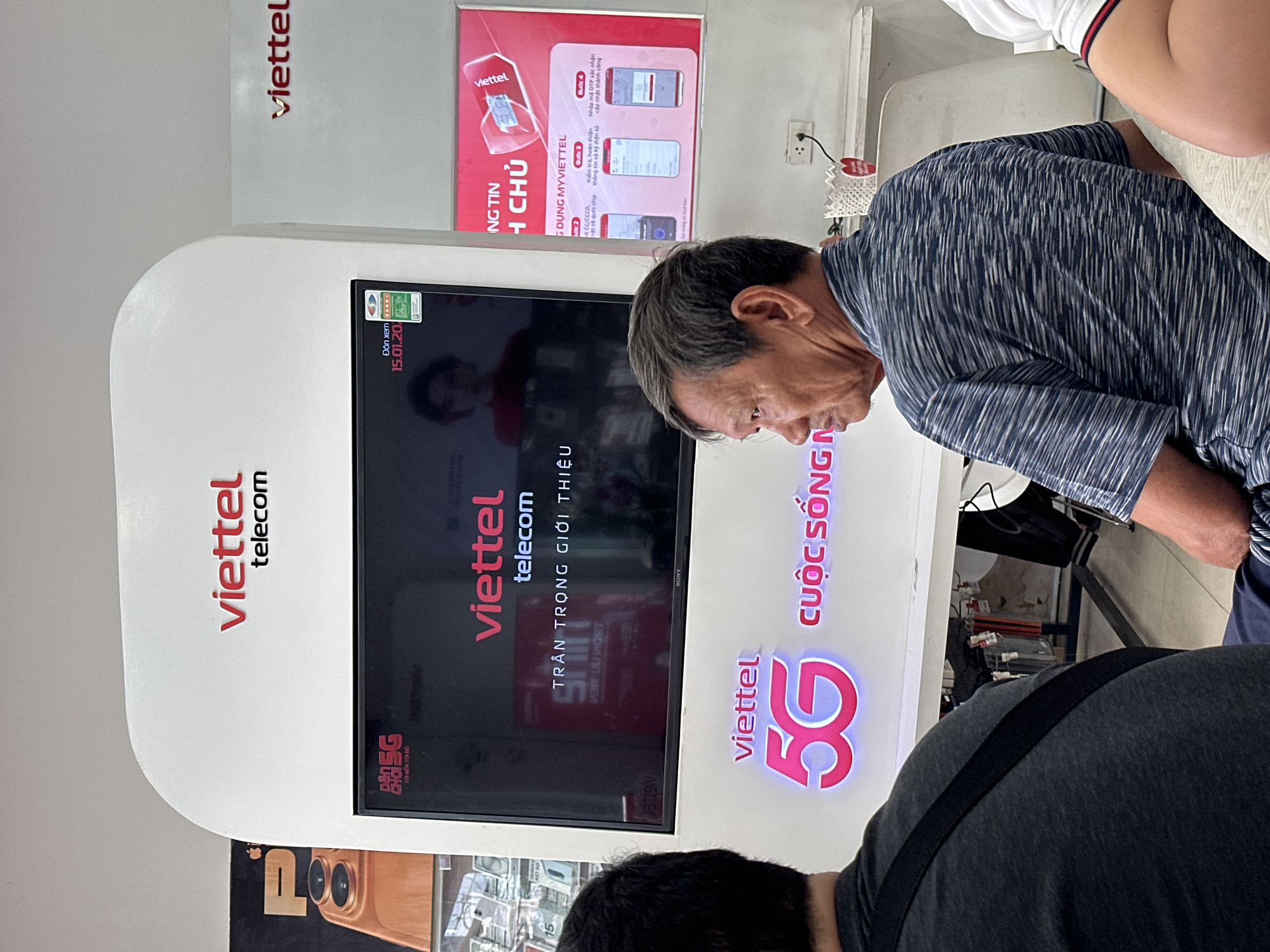 The image captures an indoor scene at a Viettel telecommunications store, likely in Nha Trang Ward, Vietnam. Two individuals are primarily visible, engaging with a modern white kiosk. In the mid-ground, an older man with short, dark hair, wearing a blue and white striped shirt, leans forward, intently looking down towards the screen embedded in the kiosk. His expression is focused. In the foreground, the back of another person is visible, wearing a dark grey t-shirt and a black strap, also appearing to look towards the kiosk. The arm of a third person in a white shirt is seen in the background.
The central feature is a sleek, white Viettel kiosk that incorporates a large, flat-screen Sony television. The screen displays "viettel telecom" branding along with the phrase "TRÂN TRỌNG GIỚI THIỆU" (Respectfully Introduces) and "Đón xem 15.01.20" (Watch on 15.01.20). To the right of the screen, the kiosk prominently features a glowing pink "viettel 5G CUỘC SỐNG" (Viettel 5G Life) logo.
On the wall above the kiosk, a framed poster provides instructions for the "MYVIETTEL" application, detailing steps such as "Bước 2" (Step 2) involving "CC/CCCD, mặt và quét chíp thông tin và ký điện tử" (ID/CCCD, face and scan chip information and e-signature) and "Bước 4" (Step 4) to "Nhập mã OTP xác nhận cập nhật thành công" (Enter OTP to confirm successful update). The "viettel telecom" logo in red is also visible on the wall. A power outlet is present on the wall. Part of another display in the bottom left shows "AS7 50" and "BSG TÔI BẾN TÔI ĐỘ". The bright, even lighting suggests the scene takes place during the daytime.