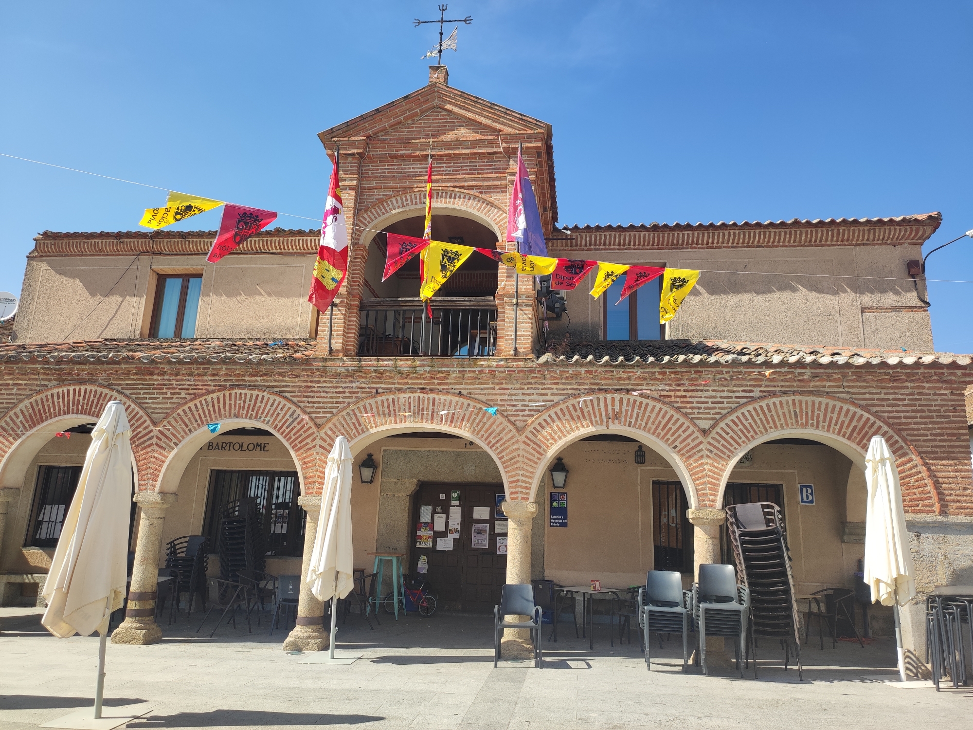 The image shows the front of a building with a brick facade and arched walkways. The building is decorated with multiple small flags strung across the front, and a larger flag hangs from a balcony. There are closed umbrellas and stacked chairs outside the building, suggesting it may be a restaurant or cafe. The sky is clear and blue, indicating a sunny day. The building appears to be located in a town square or pedestrian area, with cobblestone paving in the foreground. There are no people visible in the image. The building has a sign that reads "BARTOLOME" above one of the arched entrances. The overall scene depicts a quiet, possibly historic, location in a Spanish town, possibly Sangarcía, Spain, given the context.