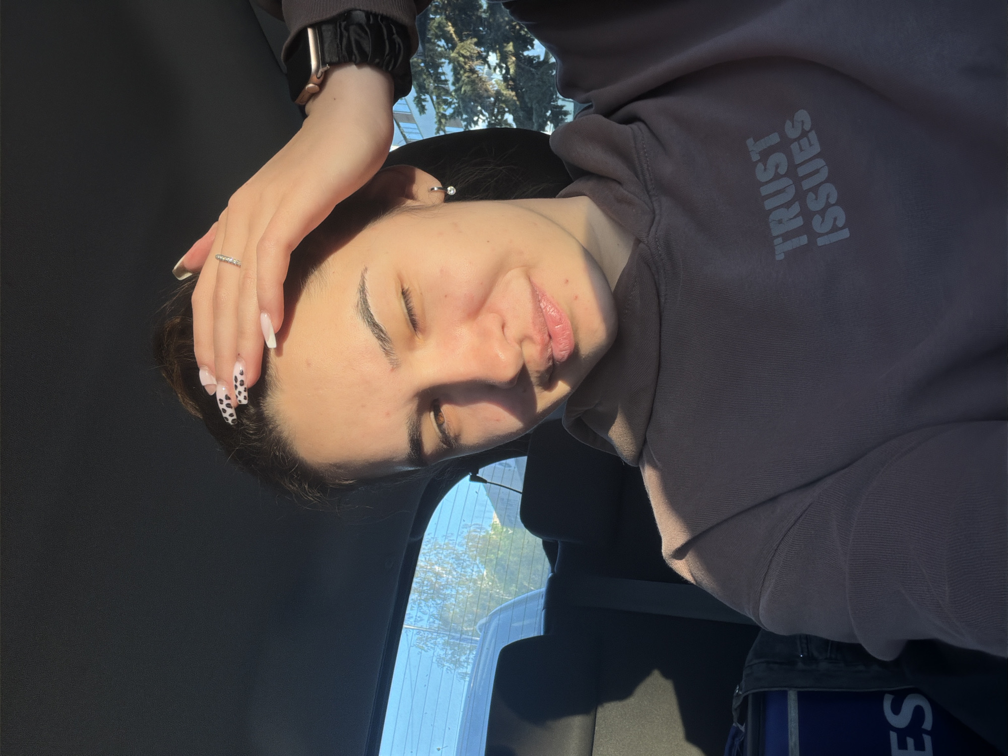 A woman with brown hair and light skin is positioned at an angle facing the camera. She is wearing a grey-brown hooded sweatshirt with the words "TRUST ISSUES" printed on the sleeve. Her left hand, adorned with a silver ring and patterned nails, rests on her head. She is wearing a watch with a black band on her left wrist. She has a small earring in her ear. The woman's eyes are partially closed. The background consists of the interior of a car, visible through the open window, and sunlight. The exact location in Sofia, Bulgaria cannot be determined from the image alone.