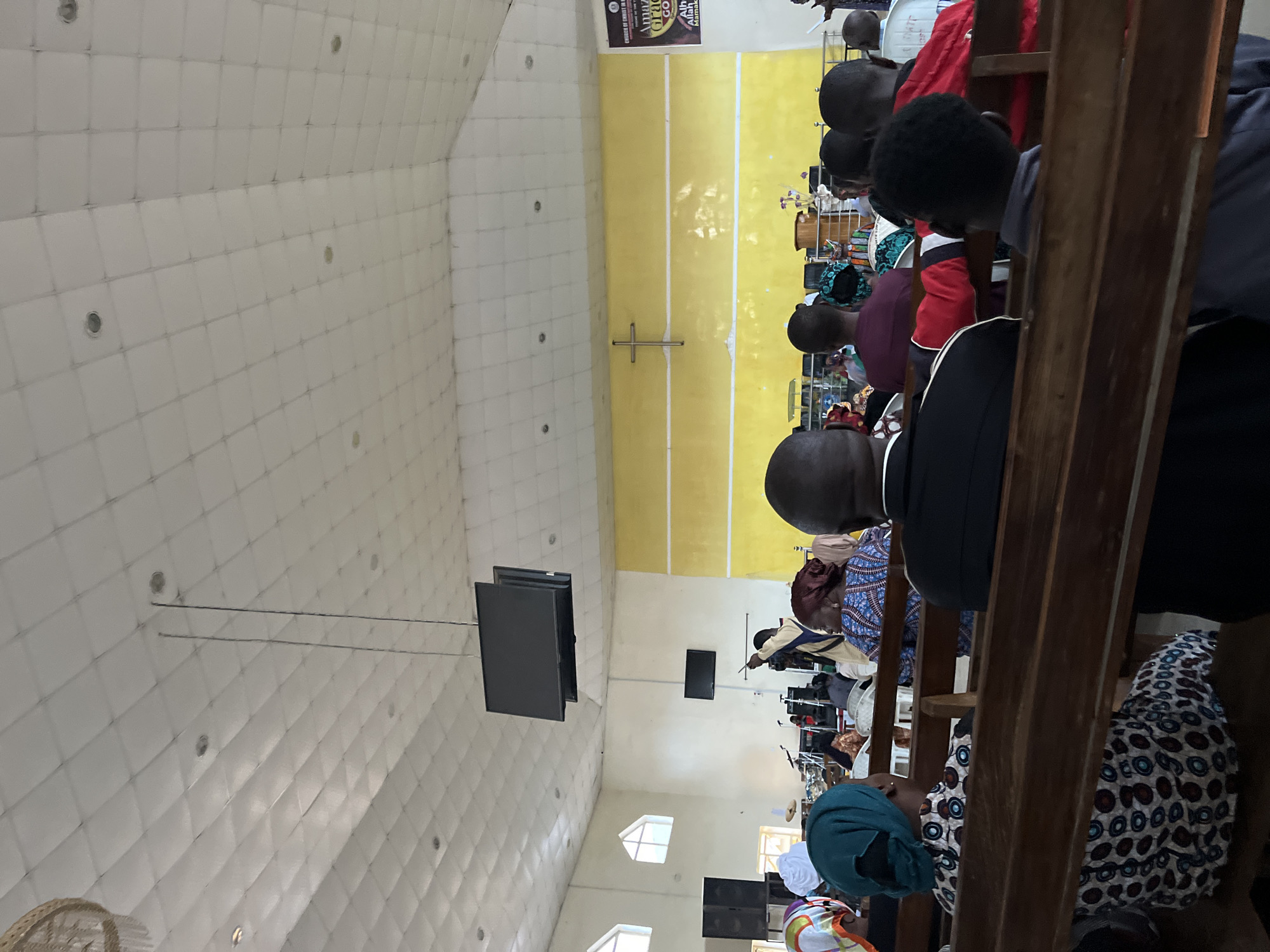The image shows the interior of a church in Jos, Nigeria. People are sitting in pews facing the front. There is a yellow-colored stage area with a cross in the middle. Screens hang from the ceiling. The ceiling is white with a grid pattern. Musical equipment is visible near the stage. Sunlight enters the church through windows.