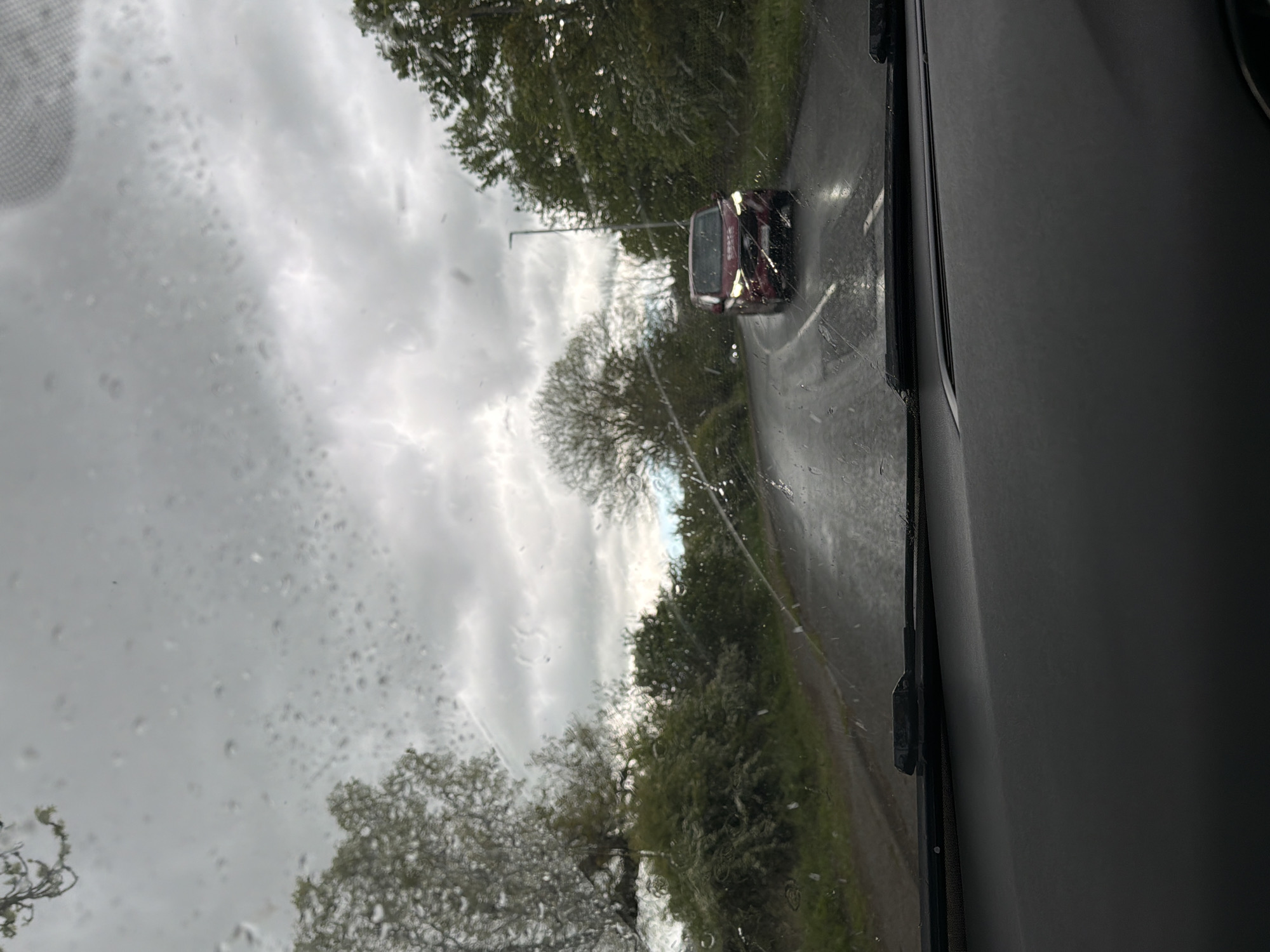 The image depicts a view from inside a vehicle, likely a car, on a rainy day in what appears to be a rural area. Through the rain-streaked windshield, a road is visible, with a red car approaching in the distance. The road is flanked by trees and greenery. The sky is overcast with heavy clouds. The interior of the vehicle is visible on the right side of the frame, showing part of the dashboard and door. There are no people visible in the scene.
Specific to the request, the location in the city of Durham, United Kingdom, cannot be specifically confirmed from the image alone; however, the rural road and vegetation are reminiscent of areas outside the city center. It would require additional information or landmarks to definitively pinpoint the location.