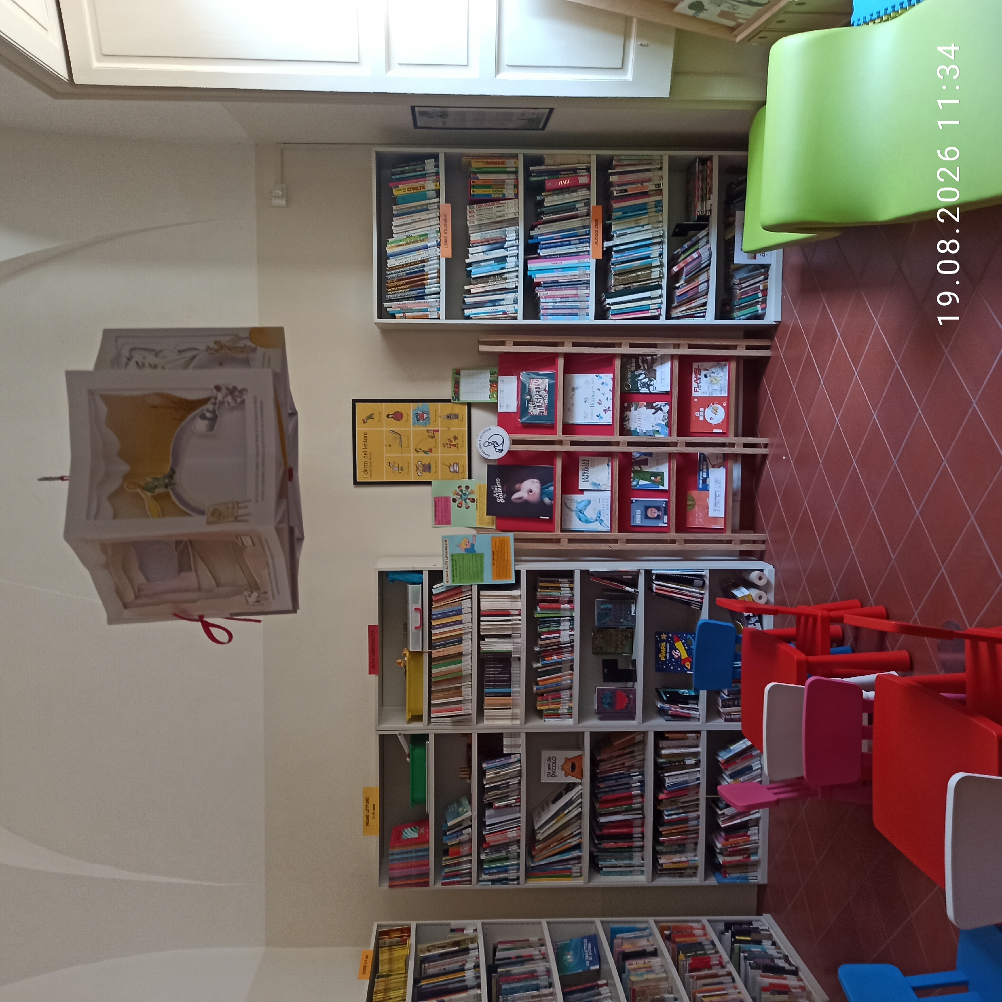 The image shows the interior of a library for children, likely in Ruvo di Puglia, Italy, indicated by the timestamp "19.08.2026 11:34" and the Italian text on signs. The scene is bright and appears well-lit, with a high, arched ceiling. The room is filled with bookshelves stocked with books. In the foreground, there are several small, colorful tables and chairs for children, made of red, blue, and pink plastic.
Dominating the center of the room is a large, cube-shaped mobile hanging from the ceiling, decorated with illustrations, likely from a children's book, depicting scenes with figures and animals. To the right of the mobile, a poster with the title "i diritti del lettore" (the rights of the reader) is displayed on the wall, accompanied by various illustrated figures and text.
The bookshelves are arranged along the walls and in freestanding units. They are organized by category, as indicated by colored labels such as "FICTION 6-9 anni" (Fiction 6-9 years old), "PRIME LETTURE 0-6 anni" (First Readings 0-6 years old), "LIBRI BILINGUE" (Bilingual Books), and "AUSOLIERE" (a category of books). Some shelves display books horizontally, while others have them vertically. Notable books visible include one with a bear cub on the cover titled "TU PICCOLO," and a book with a rocket ship on the cover titled "PIMPA."
The floor is tiled with reddish-brown square tiles arranged in a diagonal grid pattern. To the far right of the image, a bright green, undulating slide-like structure, possibly a seating or play area, is visible, partially obscuring a white, shuttered window. The overall impression is a cheerful and inviting space designed for young readers.