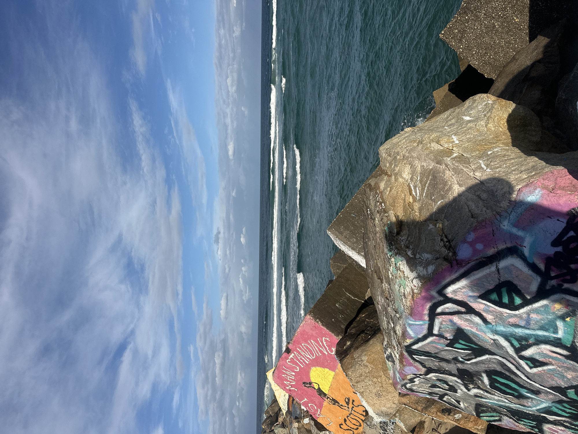 This image shows a breakwater constructed of large rocks, with graffiti visible on some of the rocks. The breakwater is located on the coast, with the ocean and waves in the background. The sky is partly cloudy. There are no people visible in the image. The graffiti includes a depiction of a person standing next to a sun, with the words "MAN STANDING" and "SCOTS" visible on a pink and orange rock face. Another rock in the foreground has colorful graffiti with black, pink, purple, and teal spray paint. The location is identified as the breakwater at the mouth of the Nambucca River in Nambucca Heads, Australia.