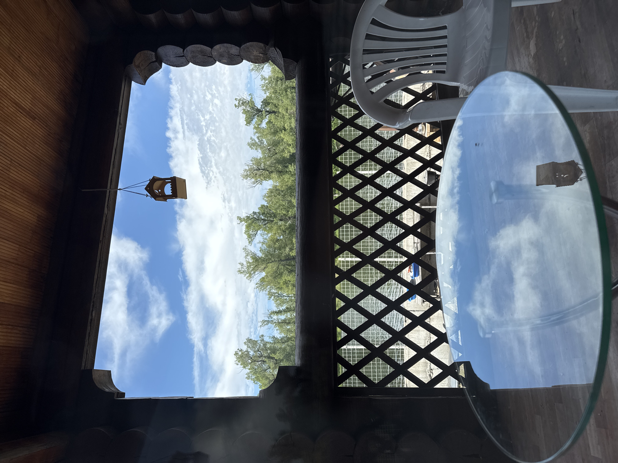 A clear view of a bright blue sky with white clouds is visible through a rustic wooden gazebo. The gazebo is constructed from dark, rounded logs and has a wooden roof. A small, wooden birdhouse hangs from a string in the center of the opening. Below the birdhouse, a decorative wooden lattice fence with diamond-shaped openings spans the width of the view. Beyond the fence, a line of green trees is visible, and further in the background, a body of water can be seen. In the foreground of the gazebo, a round glass-top table sits on the floor, reflecting the sky and the birdhouse. A white plastic chair is positioned to the right of the table. The flooring inside the gazebo appears to be wooden planks, while the floor outside the gazebo is concrete. The image has a sense of peaceful, outdoor relaxation. The location is identified as Minusinsk, Russia.
