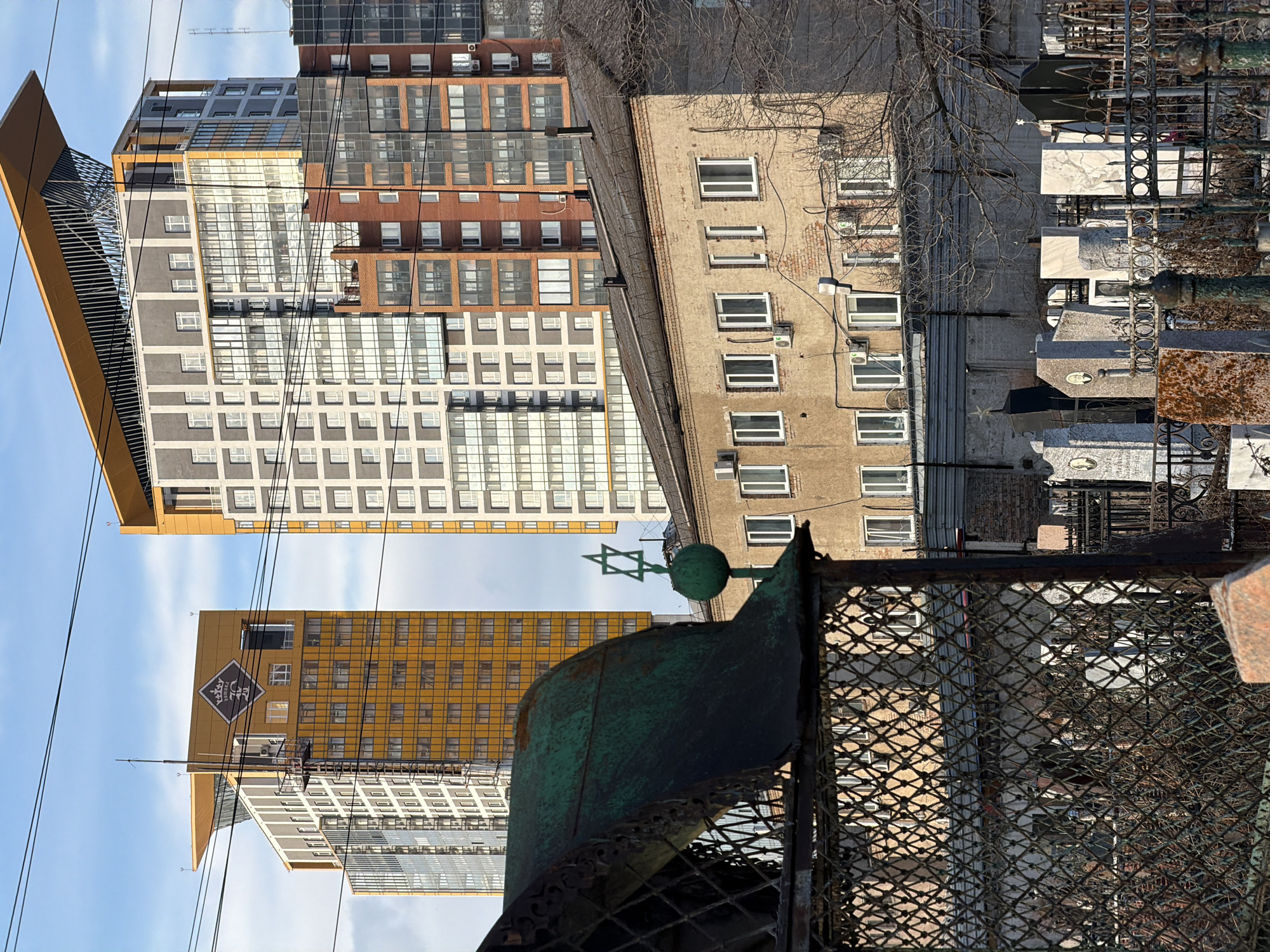 The image captures a view in Krasnoyarsk, Russia, looking out over a Jewish cemetery. The foreground consists of a weathered metal fence with a Star of David design. Beyond the fence, the cemetery contains numerous headstones and burial markers. In the background, a mix of old and modern architecture is visible, with multi-story apartment buildings including newer high-rises with distinct yellow and gray facades. The sky is light blue with some clouds. Power lines intersect the view. The overall scene depicts a juxtaposition of historical religious burial ground and modern urban development.