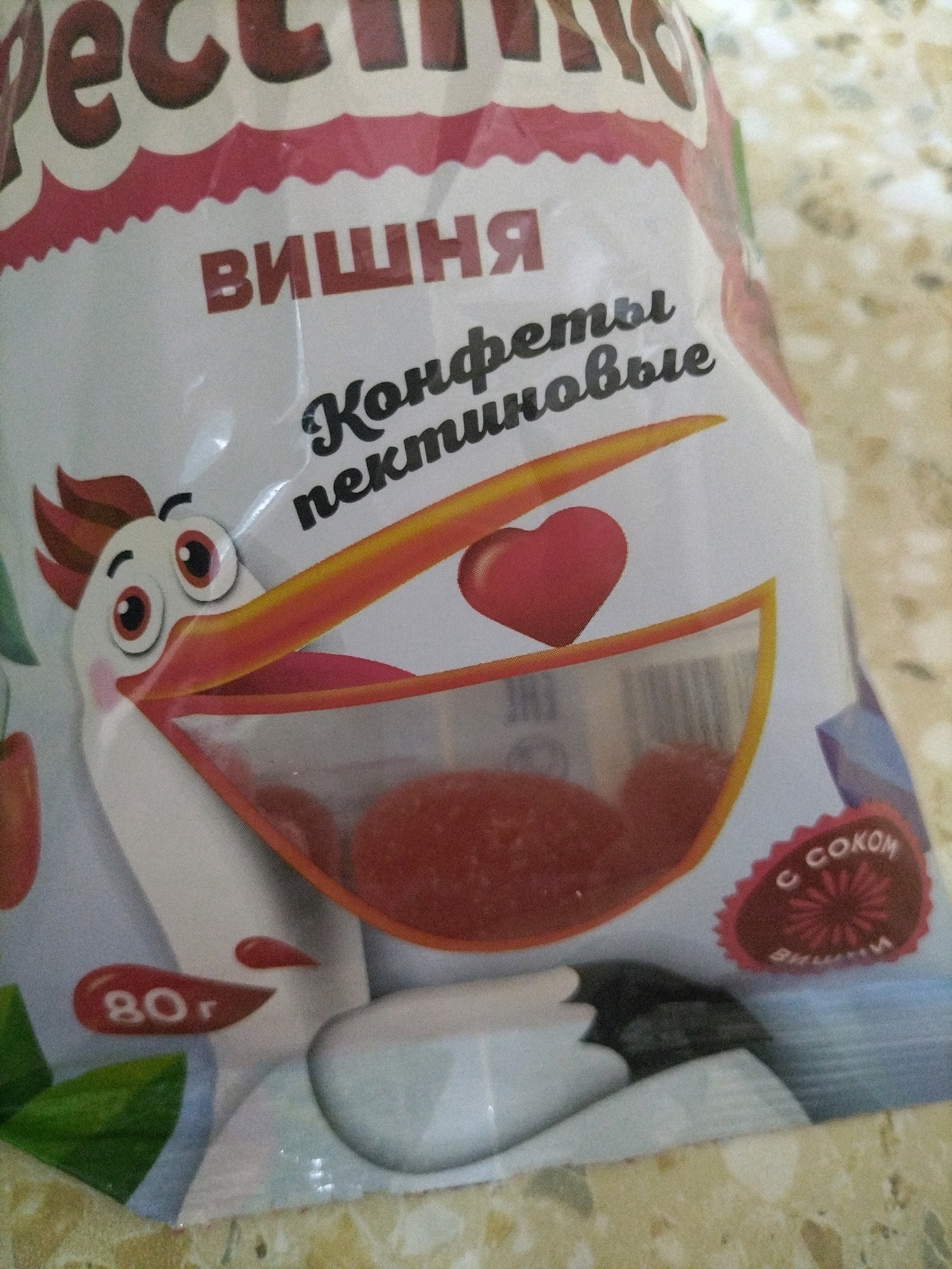 The image displays a close-up of a product package, likely for candy, with Russian text and cartoon imagery. The prominent colors are white, red, and yellow.
On the upper portion of the package, large, bold, dark red Cyrillic letters spell out "ПЕКТИН." Below this, smaller red Cyrillic letters read "ВИШНЯ" (cherry). Further down, in stylized black font, is "Конфеты пектиновые" (pectin candies).
The central and most visually striking element is a cartoon depiction of a white bird, possibly a stork or pelican, with a large orange beak and red accents around its eyes. This bird's beak is open, revealing a transparent section of the package where red, translucent candies are visible. A red heart shape is also depicted within this transparent section.
To the lower left of the bird, a red oval shape contains the white text "80 г" (80 grams). To the lower right, a red circular emblem with a stylized asterisk-like design contains the white Cyrillic text "С СОКОМ" (with juice) and below that "ВИШНИ" (cherry).
The background appears to be a textured surface, possibly a table, with a mottled beige and yellow pattern. The lighting suggests it is an indoor setting. The image quality is slightly grainy.
There is no discernible location in the city of Hrodna, Belarus, associated with this image.