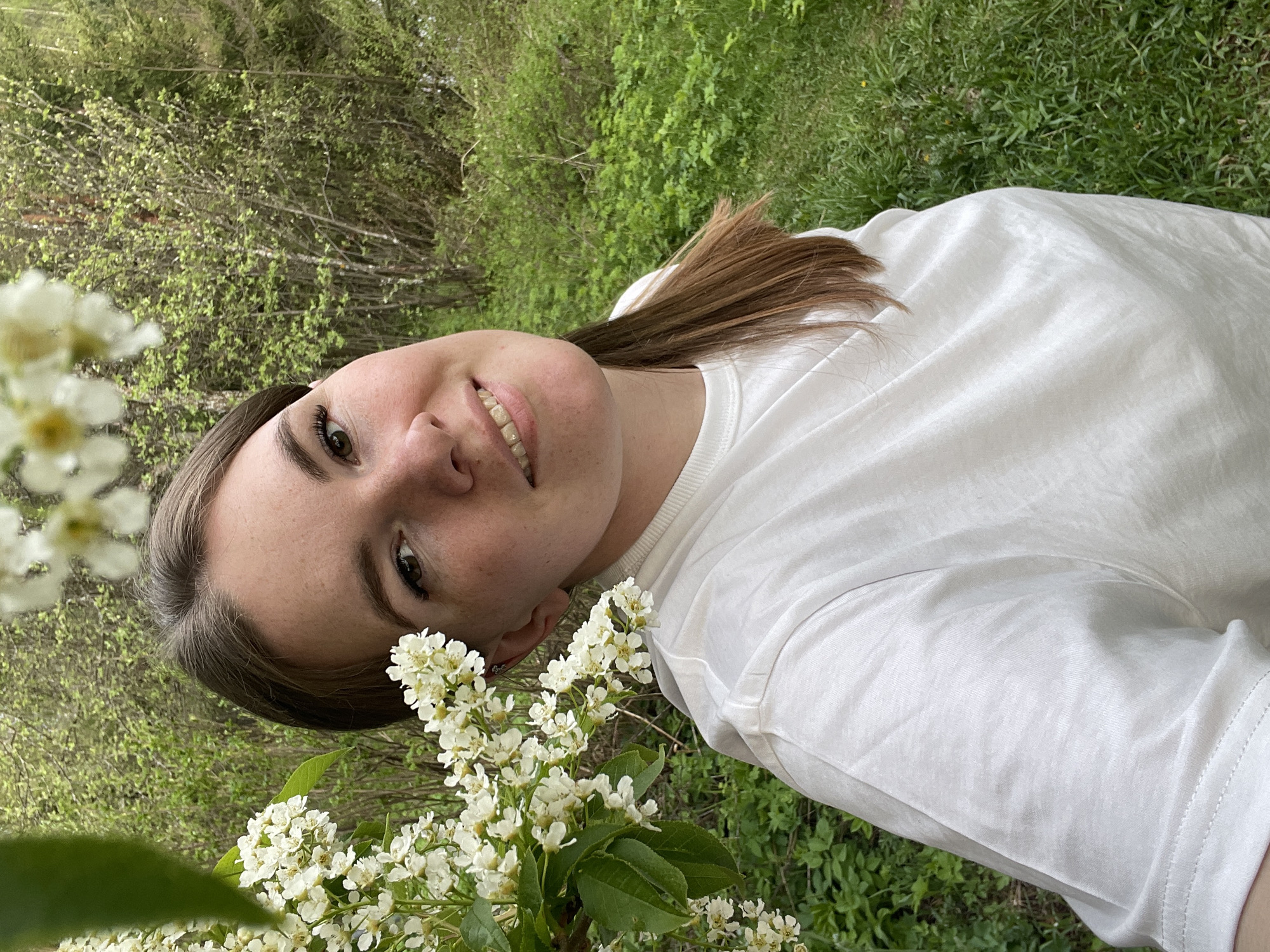 The image shows a young woman wearing a white t-shirt, with brown hair pulled back. She is smiling and looking directly at the camera. White flowers are in the foreground. The background features green foliage, likely trees and grass. The location, Zhigalki, Russia, cannot be verified from the visual information provided.