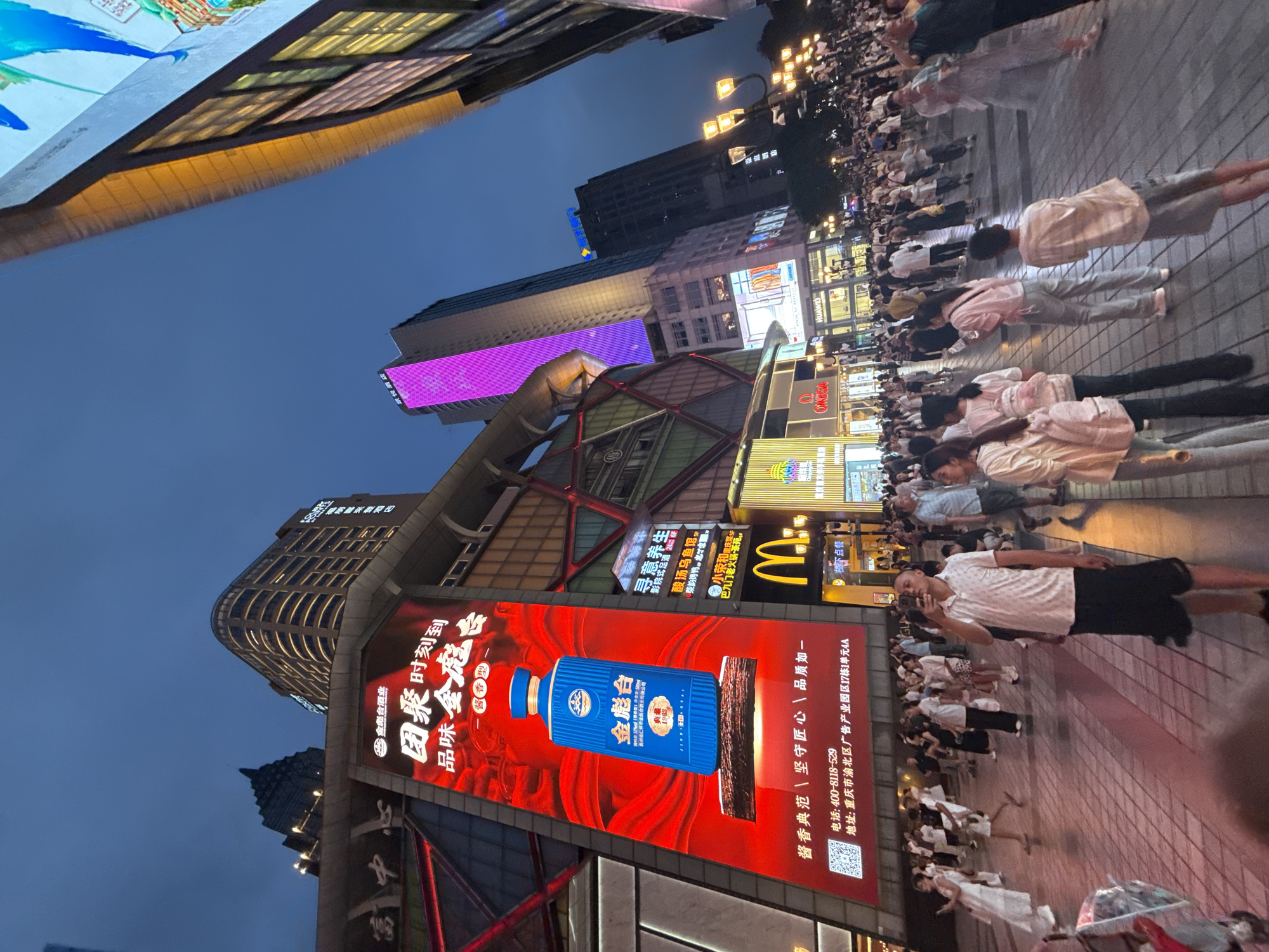 The image captures a bustling street scene in Yuzhong District, China, at dusk. Towering buildings with illuminated signage dominate the skyline, the most prominent being a large, bright red billboard displaying an advertisement for "Jingtai" (金酱台) liquor. The advertisement features a blue bottle of the liquor against a red background with Chinese text. To the left of the billboard, a building facade features geometric patterns with red and white lights.
Below the billboard, a crowd of people is walking on a paved walkway. Many are looking at their phones or engaging in conversations. Among the crowd, a man in a white polo shirt and black shorts is taking a photo with his phone. Several women are walking in groups, some carrying bags and backpacks. Commercial establishments with brightly lit storefronts, including a McDonald's, are visible. Streetlights illuminate the scene, and the sky is a deep blue, indicating the transition from day to night. The overall impression is one of a lively urban environment with a mix of commercial activity and pedestrian traffic.