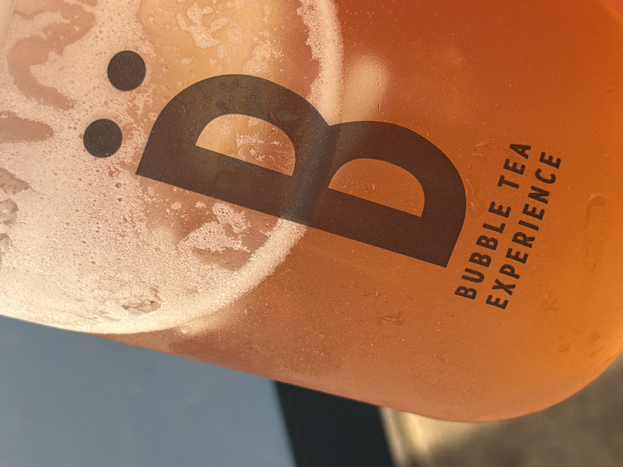 A close-up, slightly angled view of a clear plastic cup filled with a beverage, likely a bubble tea. The liquid inside appears orange. Condensation is visible on the exterior of the cup, with bubbles and frost-like textures near the top. The cup's branding includes a large, stylized "B" logo in black, and the words "BUBBLE TEA EXPERIENCE" printed vertically in black block letters. The cup rests on a dark surface. Based on the brand and available information, the location is likely Rishon LeZion, Israel.