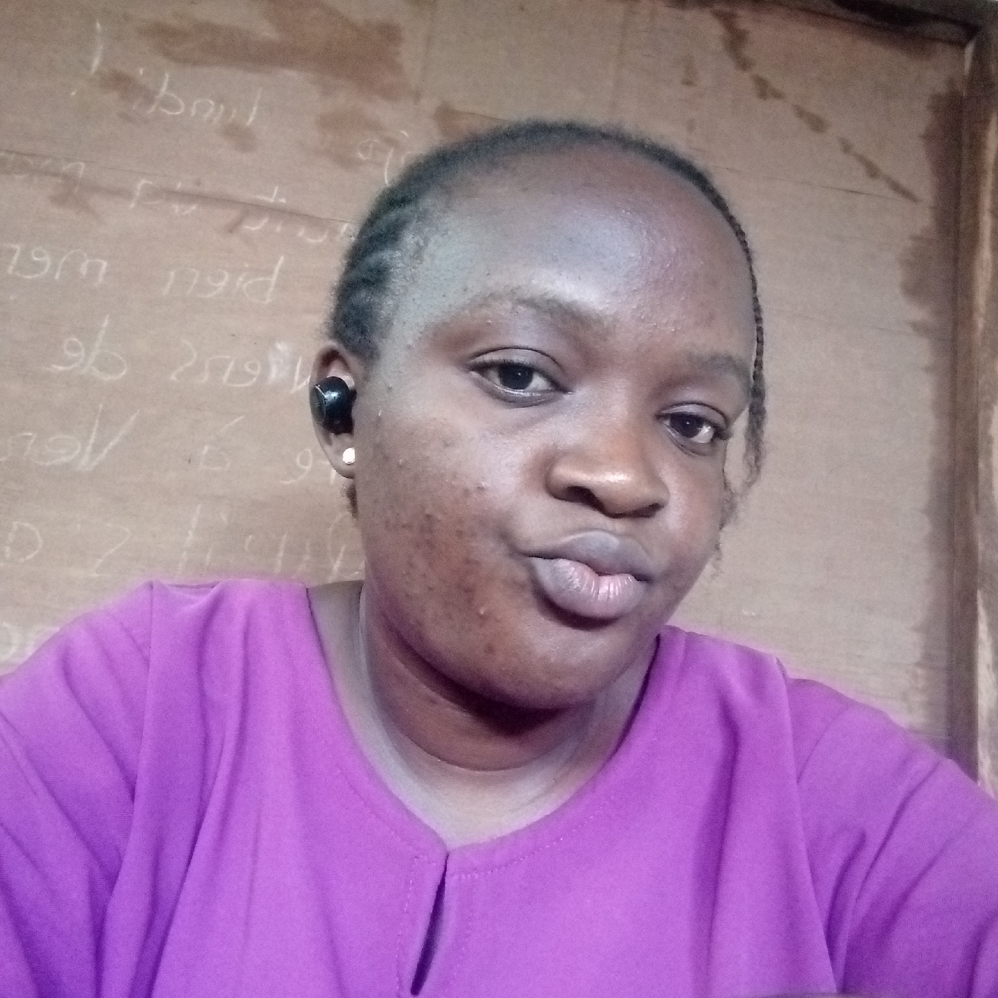 A medium shot shows a woman with brown skin, wearing a magenta blouse. She is making a kissing face towards the camera. She has short braids. She has a black wireless earbud in her left ear and a silver stud earring in the same ear. The background is a tan wall with faint writing on it. The location in Ukwu-Abwa, Nigeria is unknown.
