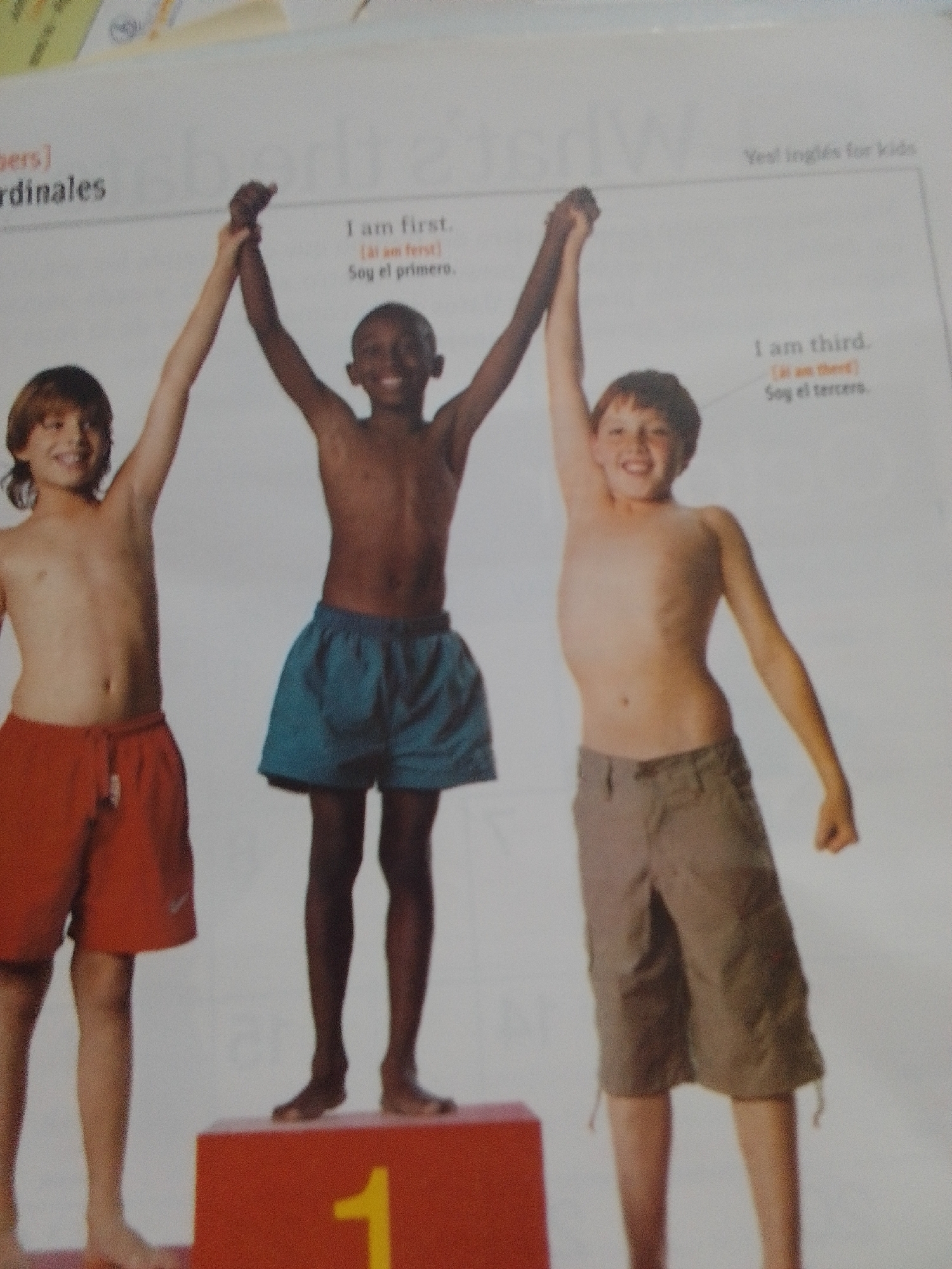 This media file shows three young boys on a podium. The boy on the left is wearing red shorts and has light brown hair. The middle boy is wearing blue shorts and has dark skin. The boy on the right is wearing khaki shorts and has light brown hair. All three boys have their arms raised, with the two boys on the ends holding hands above the middle boy's head. The podium is red, and the number "1" is visible on the front of it in yellow. Text above the boys indicates they are participating in some kind of competition where they are being recognized for their placement. The text reads "I am first," "Soy el primero," "I am third," and "Soy el tercero." There is also text in the upper right corner that says "Yes! inglés for kids," suggesting this is an educational context. The scene appears to be indoors, likely a photo shoot for a book or educational material. The boys have happy expressions, suggesting they are pleased with their accomplishments.