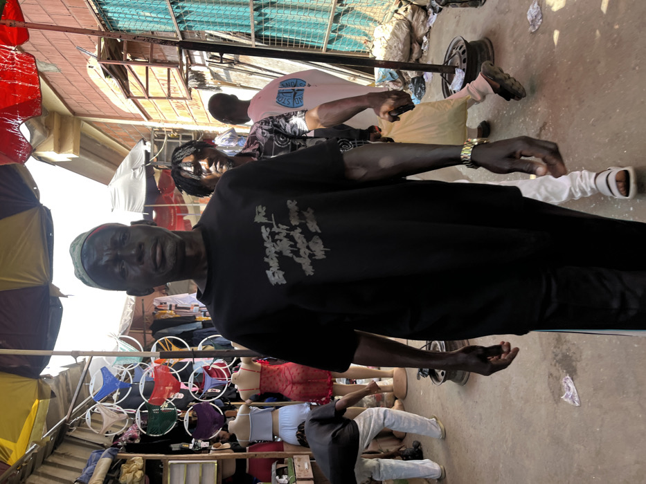 This image depicts a bustling outdoor market scene in Abuja, Nigeria, likely an informal street market.
In the foreground, a dark-skinned adult male stands centrally, facing the camera. He wears a black short-sleeved t-shirt with the light-colored text "Embrace The MIGHT Unleash Your WILD SOUL" printed on the chest. He has dark trousers with a light stripe and wears a patterned green cap. A gold-colored watch or bracelet is visible on his left wrist. His skin exhibits a reflective sheen.
Behind him and slightly to the right, another dark-skinned adult male with dreadlocks walks forward, also looking towards the camera. He wears a dark, patterned short-sleeved shirt and carries a light-colored plastic bag in his right hand. A gold-colored watch is visible on his left wrist. Further back on the right, a lighter-skinned male in a light pink t-shirt with a blue design on the back walks away from the camera. To the far left, another individual in a dark shirt and light trousers is bent over, seemingly attending to items on the ground near several mannequins.
The market environment features multiple stalls. On the left, a stall displays various undergarments, including colored panties on circular hangers and lingerie on mannequins (a light blue two-piece set and a sheer red teddy). Stacked clothing or fabric is visible on shelves. Overhead, large market umbrellas, one striped red and yellow, provide shade. The ground is a worn, light-colored surface, possibly concrete or packed earth, with scattered debris including a crumpled plastic bag and discarded metallic objects resembling tire rims near a support pole on the right. In the background on the right, a light-colored building with a turquoise metal gate or screen is visible. The scene is illuminated by bright daylight.