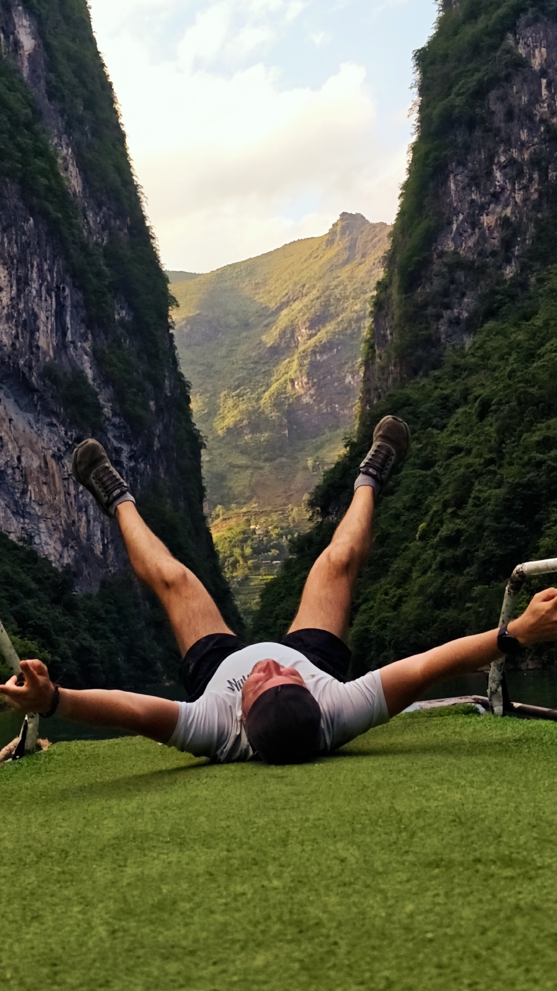 A man lies supine on a green surface, legs raised, arms extended. The scene is framed by steep, tree-covered cliffs typical of the Đồng Văn Commune in Vietnam. A landscape with verdant hills and a small settlement is visible in the distance between the cliffs, beneath a partly cloudy sky. The man wears dark shorts, a white t-shirt, shoes with dark socks, and a black wristband. He appears relaxed, with his arms outstretched. A metallic bar and rope are near his hands.