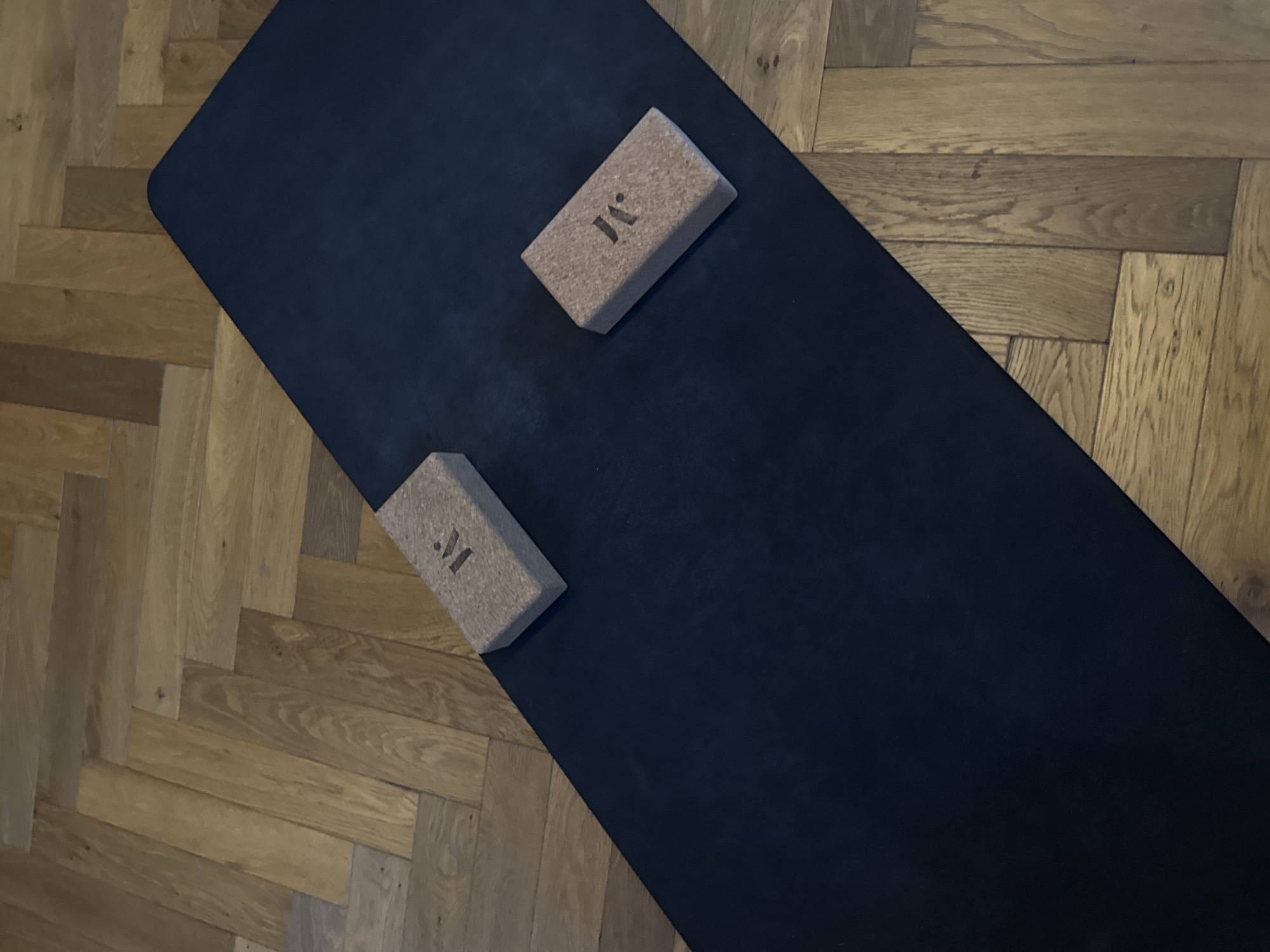 A still-life photograph depicts a black yoga mat resting on a parquet wood floor. Two rectangular cork yoga blocks, each bearing a stylized "M" logo, are positioned on the mat, one towards the top right and the other towards the bottom left. The setting suggests a personal workout space, possibly within a home. There are no people visible in the frame. The precise location within London, United Kingdom cannot be determined from this image alone.