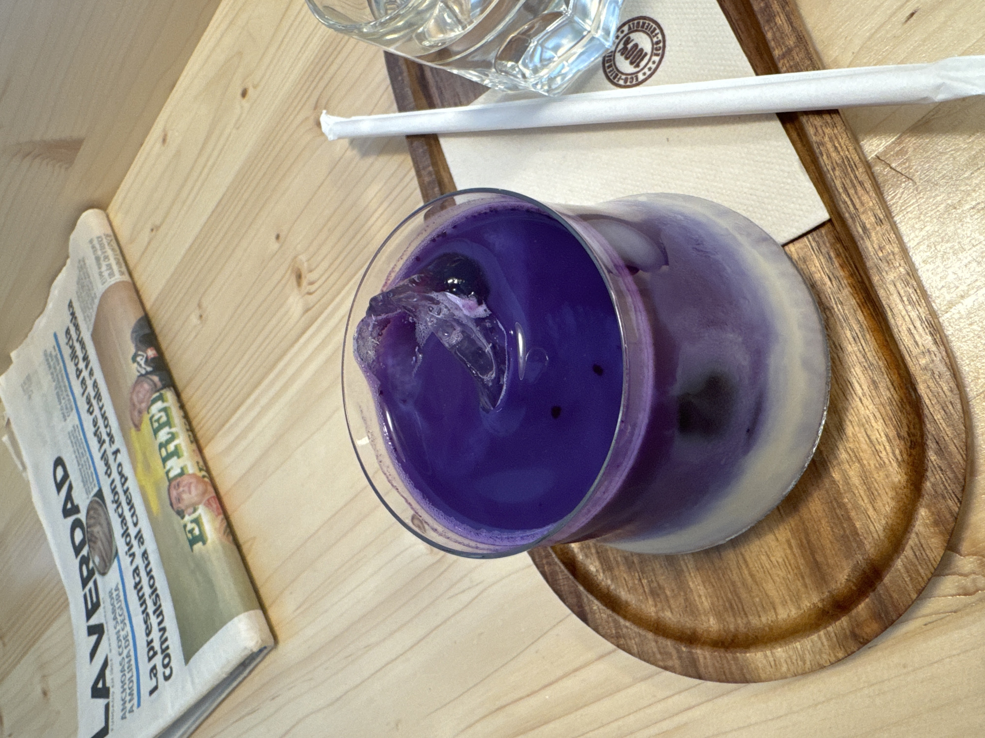 The image depicts a close-up, high-angle view of a purple beverage in a glass with ice, placed on a wooden coaster. The coaster rests on a light-colored wooden table. To the right of the beverage, a white paper-wrapped straw and a glass of water are visible. Behind the beverage and coaster, a newspaper with Spanish text, including the headline "LA VERDAD," is partially visible. A small section of a logo with "100%" is visible on the napkin under the glass of water. The environment appears to be an indoor setting, possibly a cafe or restaurant. The lighting suggests it is daytime. There are no people visible in the image, and no specific details of Murcia, Spain, are discernible from the provided view.