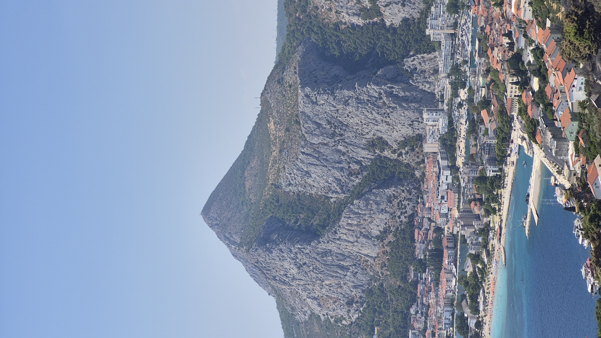 This is a high-angle, vertical, full-shot image of a coastal city dominated by a large, rocky mountain. The sky is clear and pale blue. The mountain's peak is in the upper portion of the frame, and it's covered in sparse vegetation and large rock formations. The city stretches along the coastline at the base of the mountain.
The city features a variety of buildings, including residential structures with red-tiled roofs, and taller, modern buildings. There is a beach area with numerous sunbathers visible. The ocean in front of the city is a vibrant turquoise and deep blue, with several boats docked or anchored in a harbor. A breakwater extends into the water. Trees and greenery are scattered throughout the city and on the lower slopes of the mountain. There are also some cell towers visible on the mountain ridge. The overall impression is of a sunny day in a scenic European coastal town.